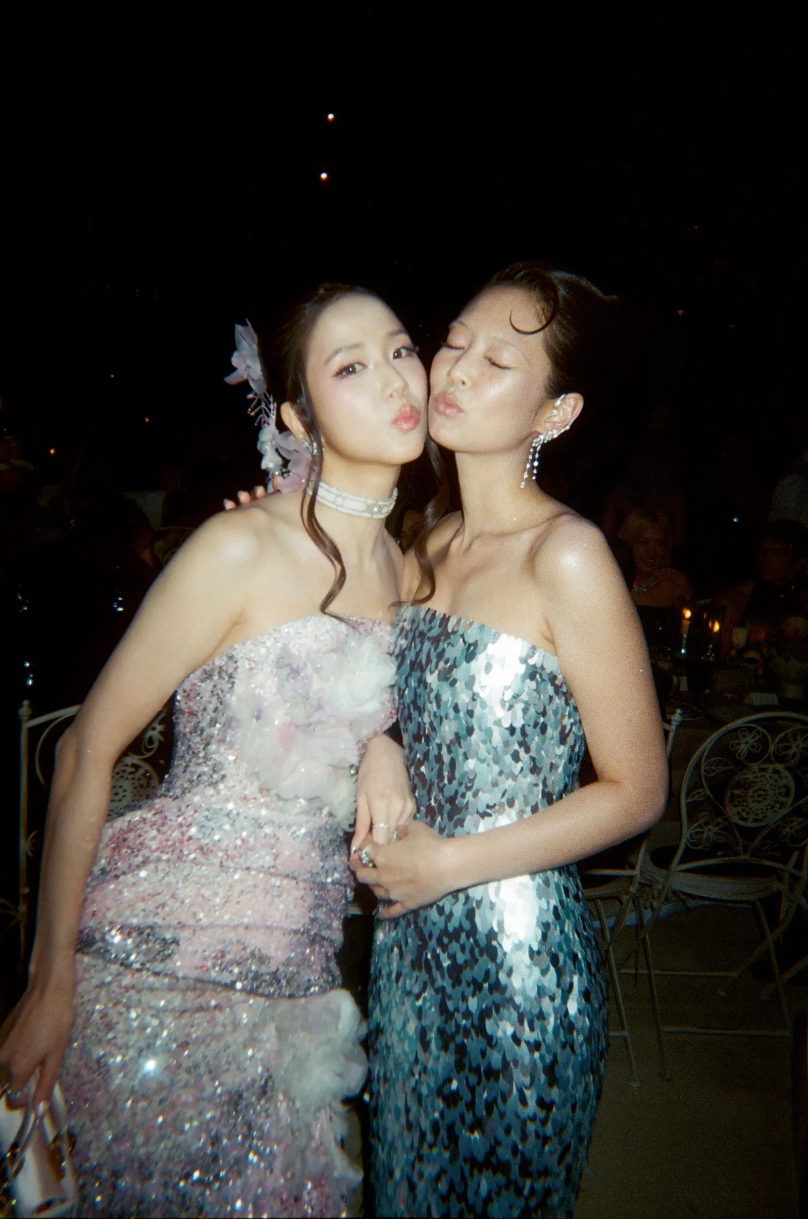 Two women in sparklingformal gowns kiss each other at a nighttime party, with one wearing a pinkish dress and the other a blue-green sequined dress.