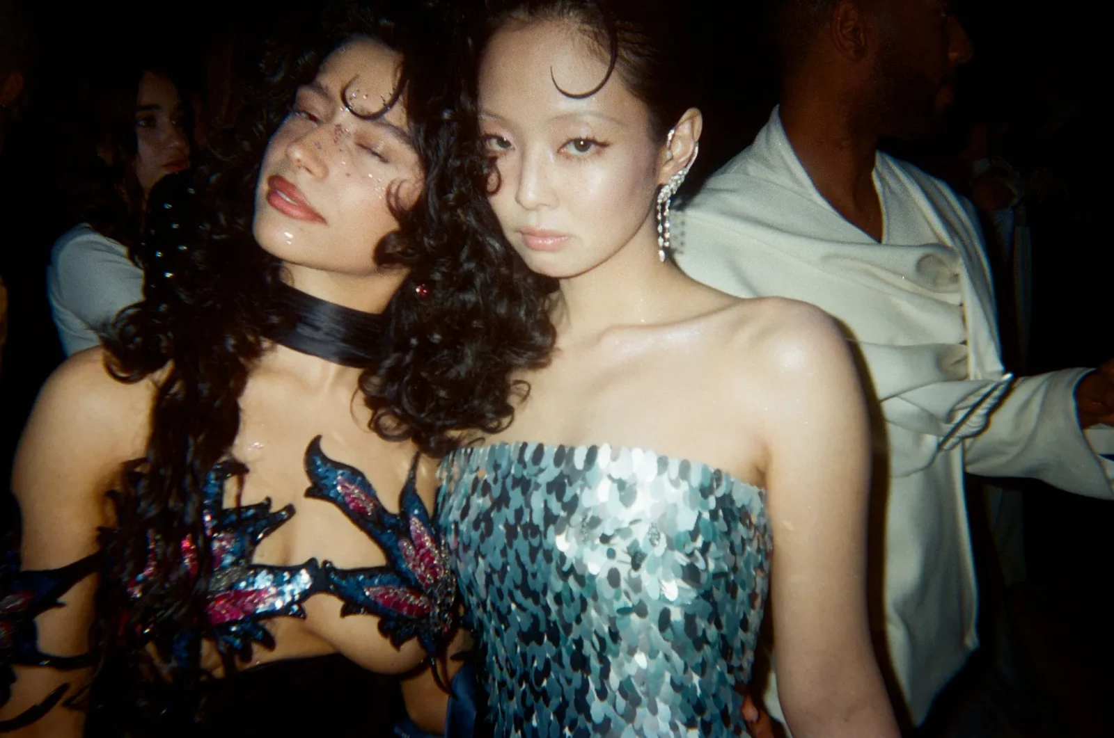 Two women posing closely at a dimly lit party; left woman with curly hair and a sequined dress has her eyes closed, while the right woman looks at the camera and wears a dangling earring.