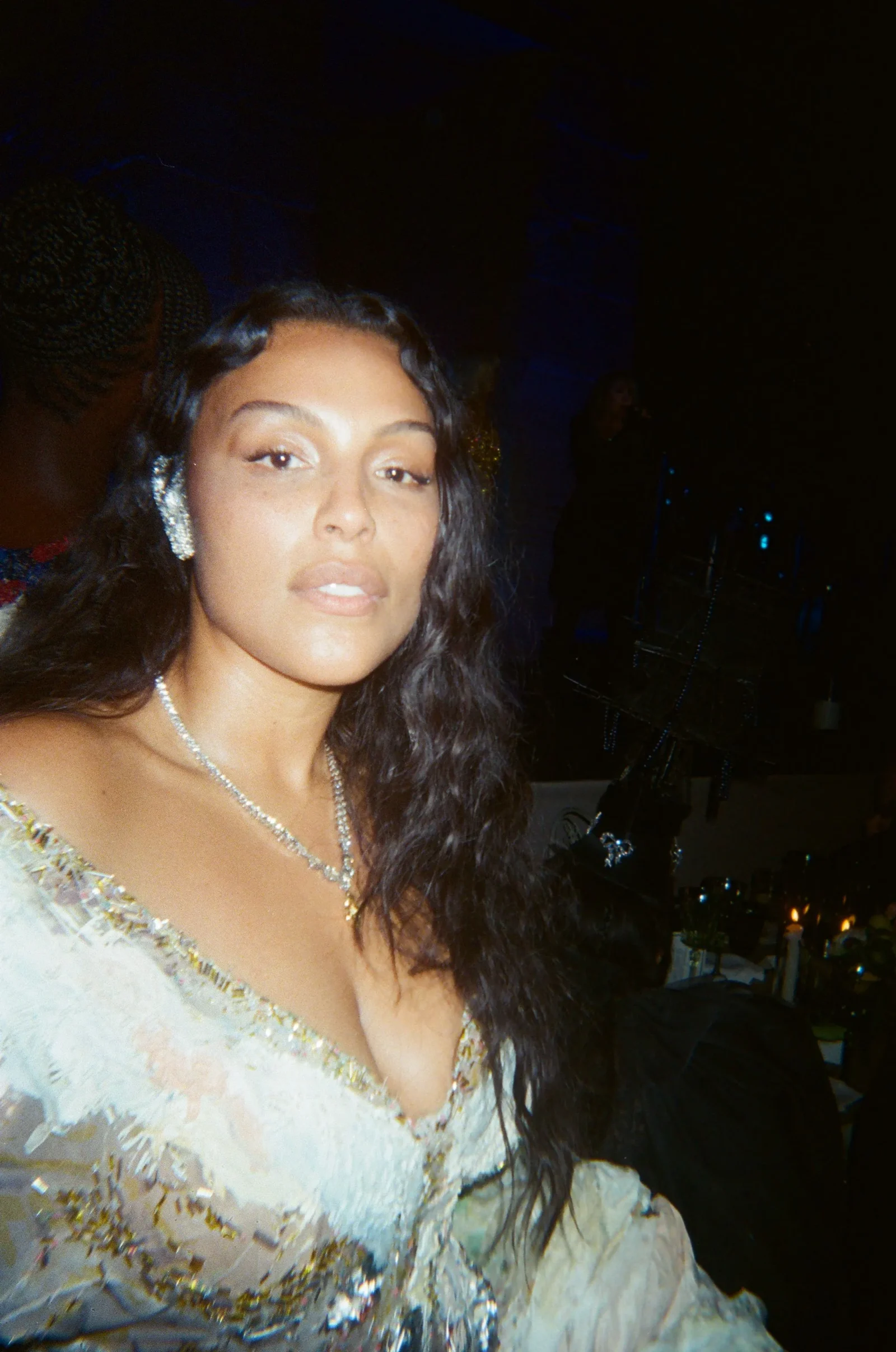 Close-up of a woman with long dark hair wearing a shimmery light-colored dress at a dim party setting, looking at the camera.