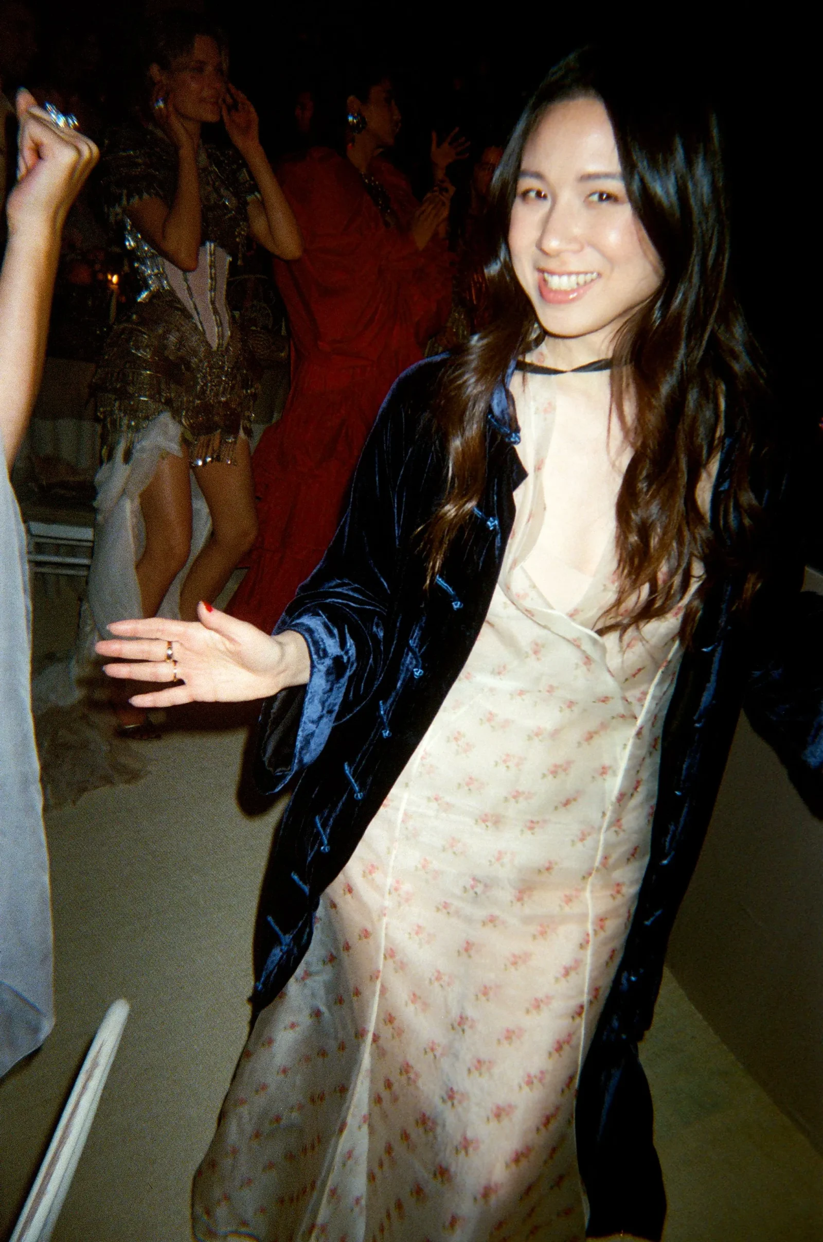 Smiling woman in a floral dress and dark velvet jacket reaching toward the camera at a party, with guests in festive outfits in the background.