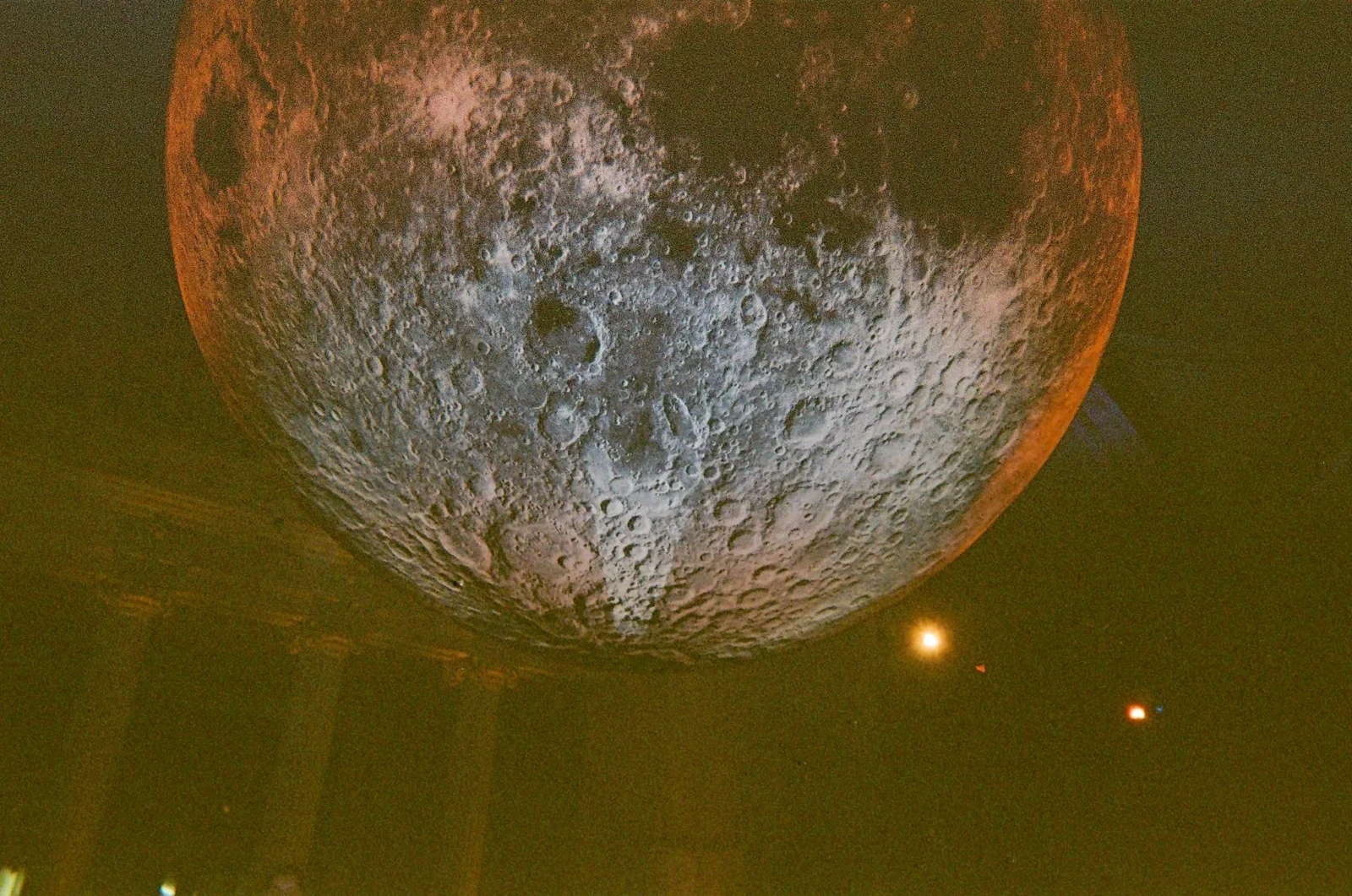 Close-up of the Moon's cratered surface with an orange-tinted glow and small lights in the foreground