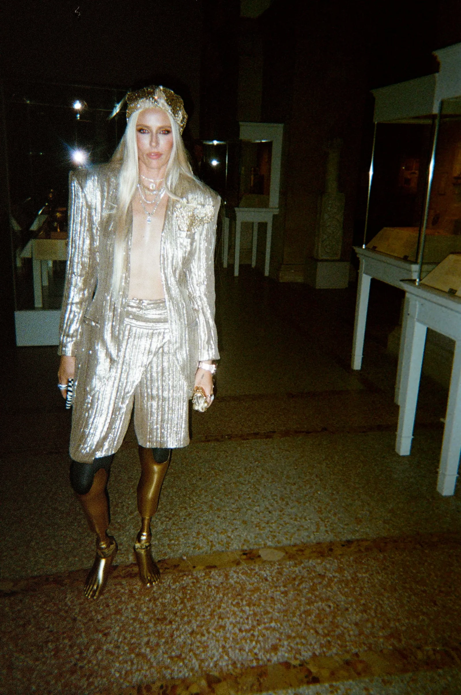 Person in a shiny silver suit with crown, long blonde hair, standing indoors wearing jewelry and gold boots.
