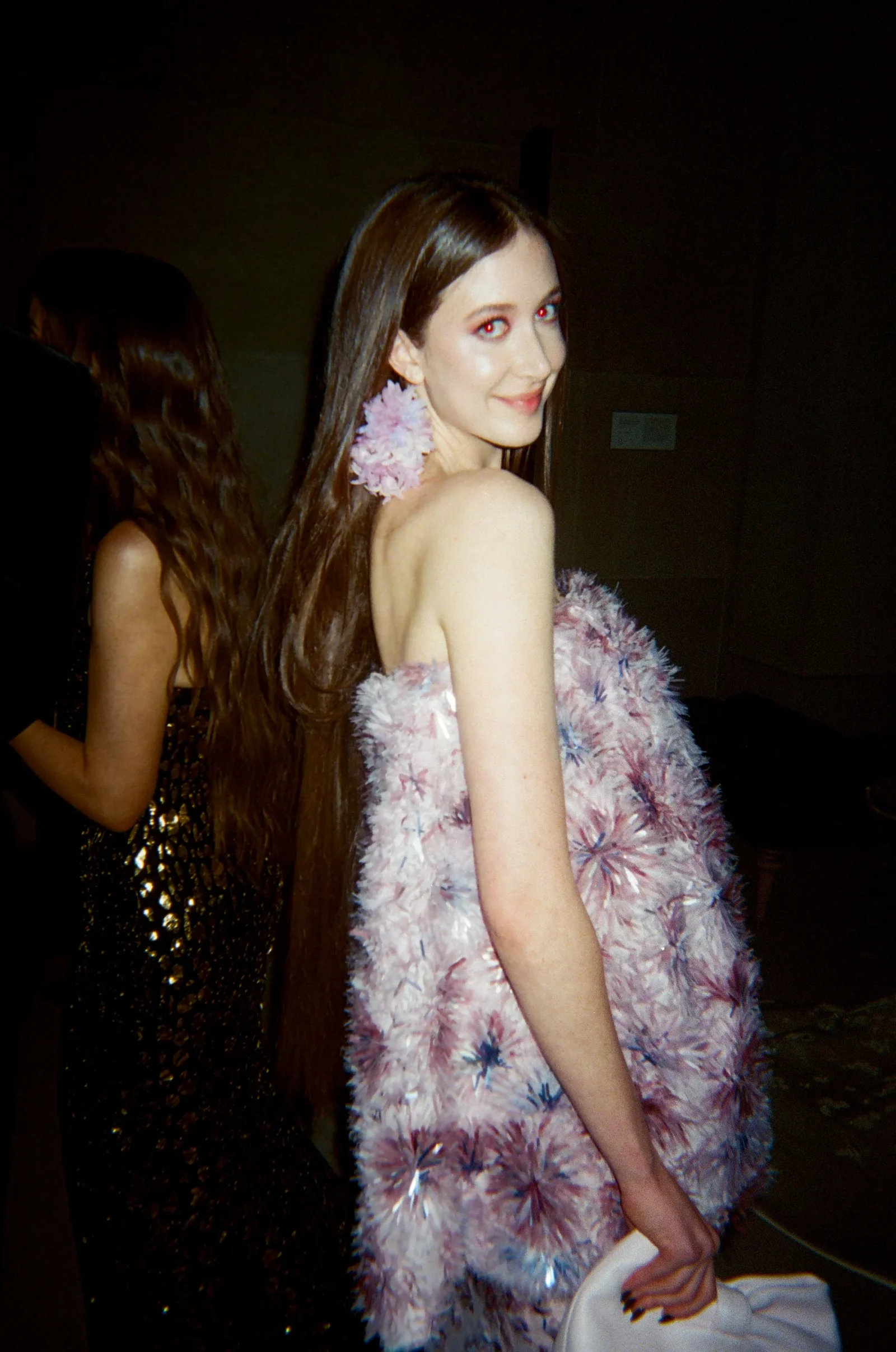 Woman with long brown hair wearing a pink feathered dress, smiling at the camera in a dim venue.