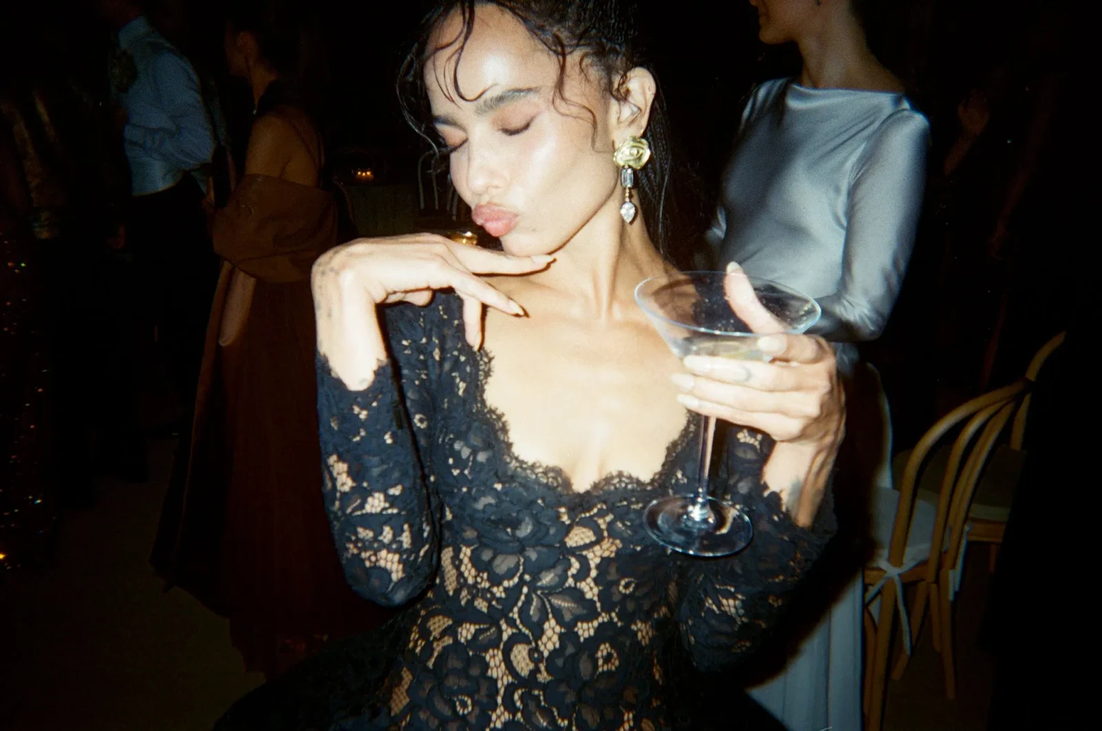 Woman in a black lace dress holding a cocktail glass with eyes closed at a dimly lit party