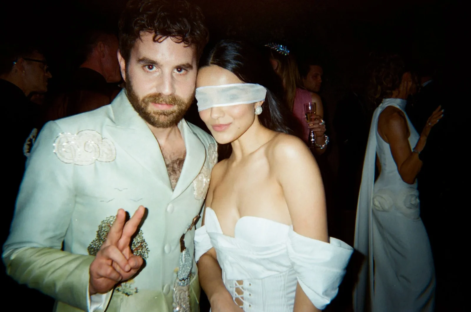 Man in a pale suit with embroidered details poses with a woman in a white off-shoulder dress who is blindfolded with a white ribbon at a crowded party or club.
