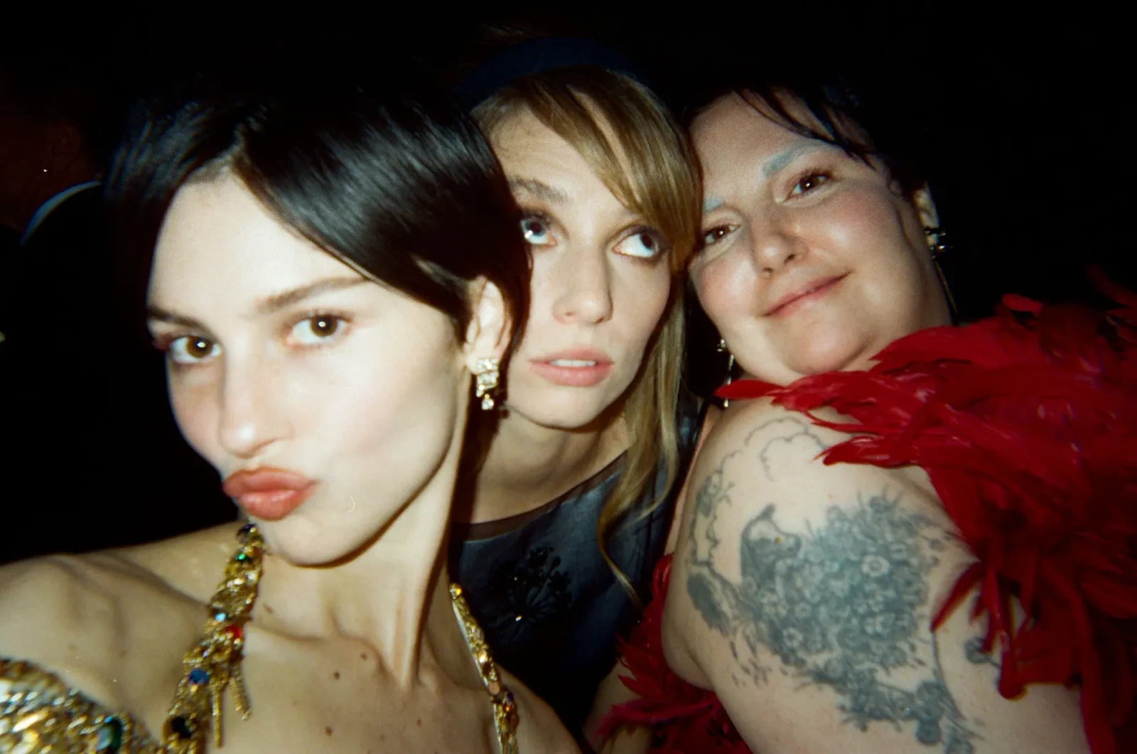 Three women pose for a close selfie at a dim party, smiling with a dark background behind them.