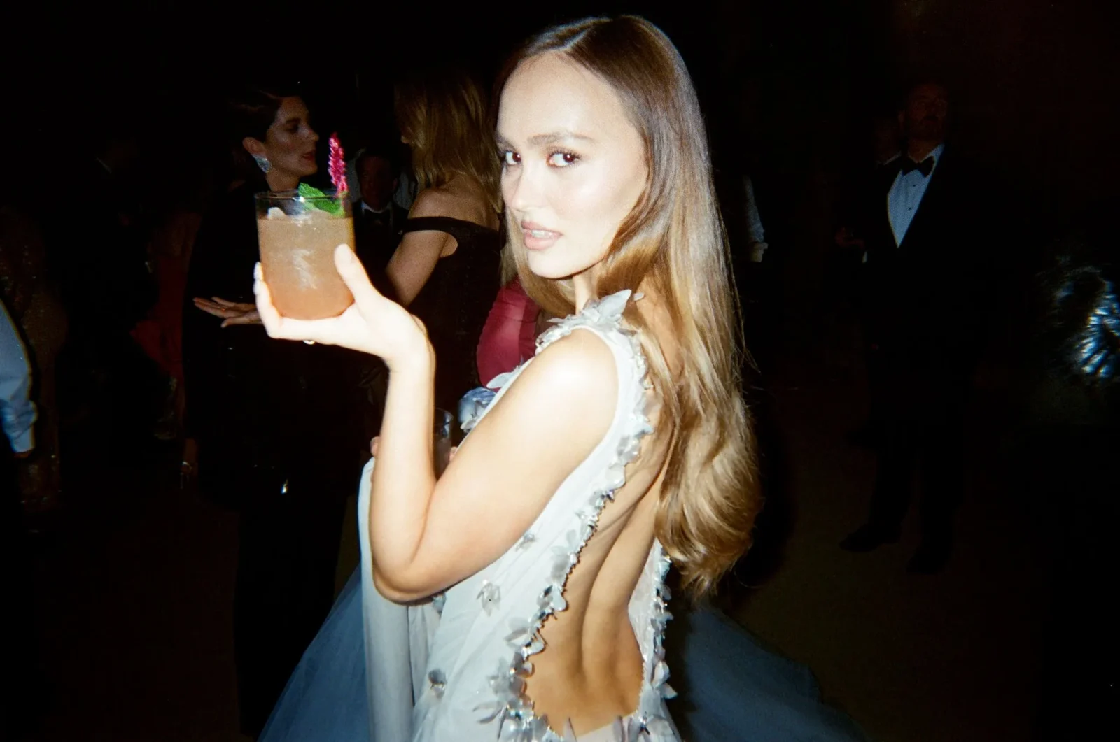 Woman in a light blue, floral-trim dress at a party holding a cocktail with a pink garnish and straw, smiling at the camera.