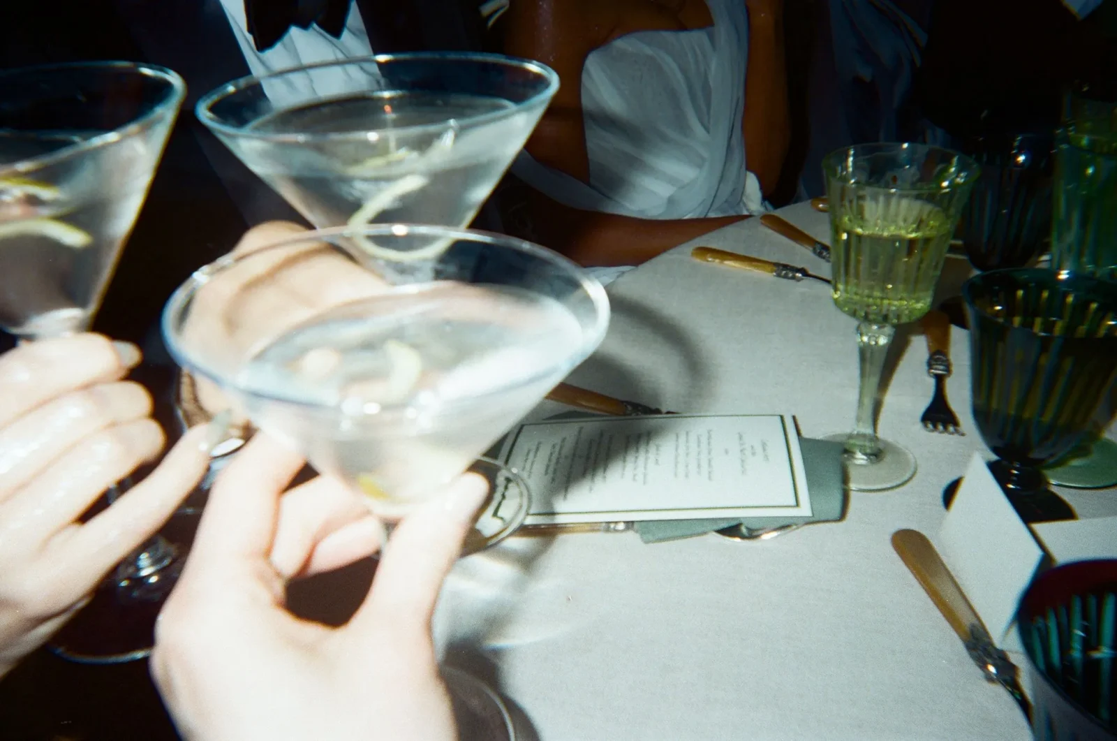 Close-up of hands holding cocktail glasses raised for a toast at a dimly lit dinner, with menus and utensils on a white tablecloth.