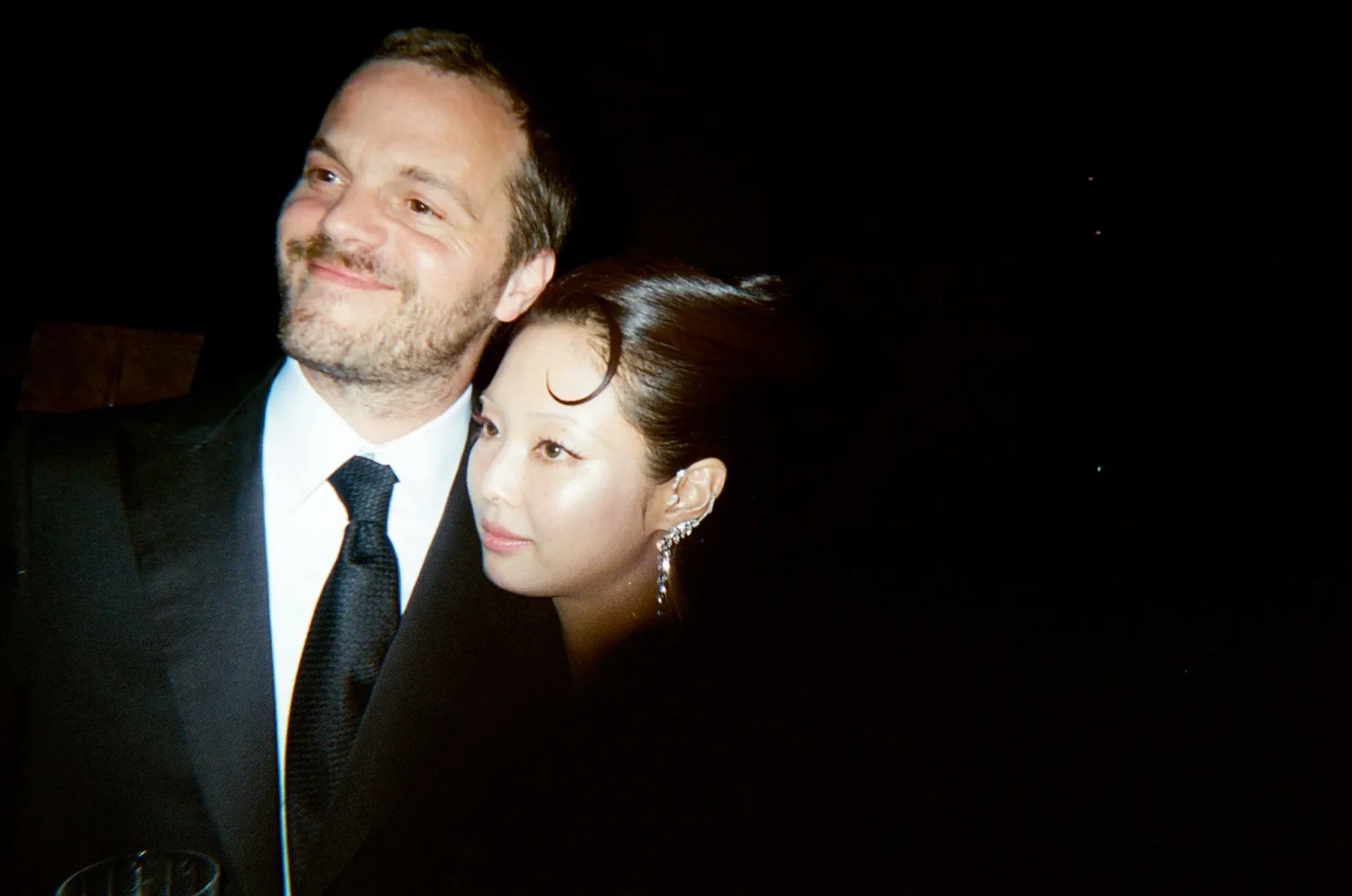 Man in a suit and tie smiles as a woman rests her head near his shoulder in a dark, night-time setting