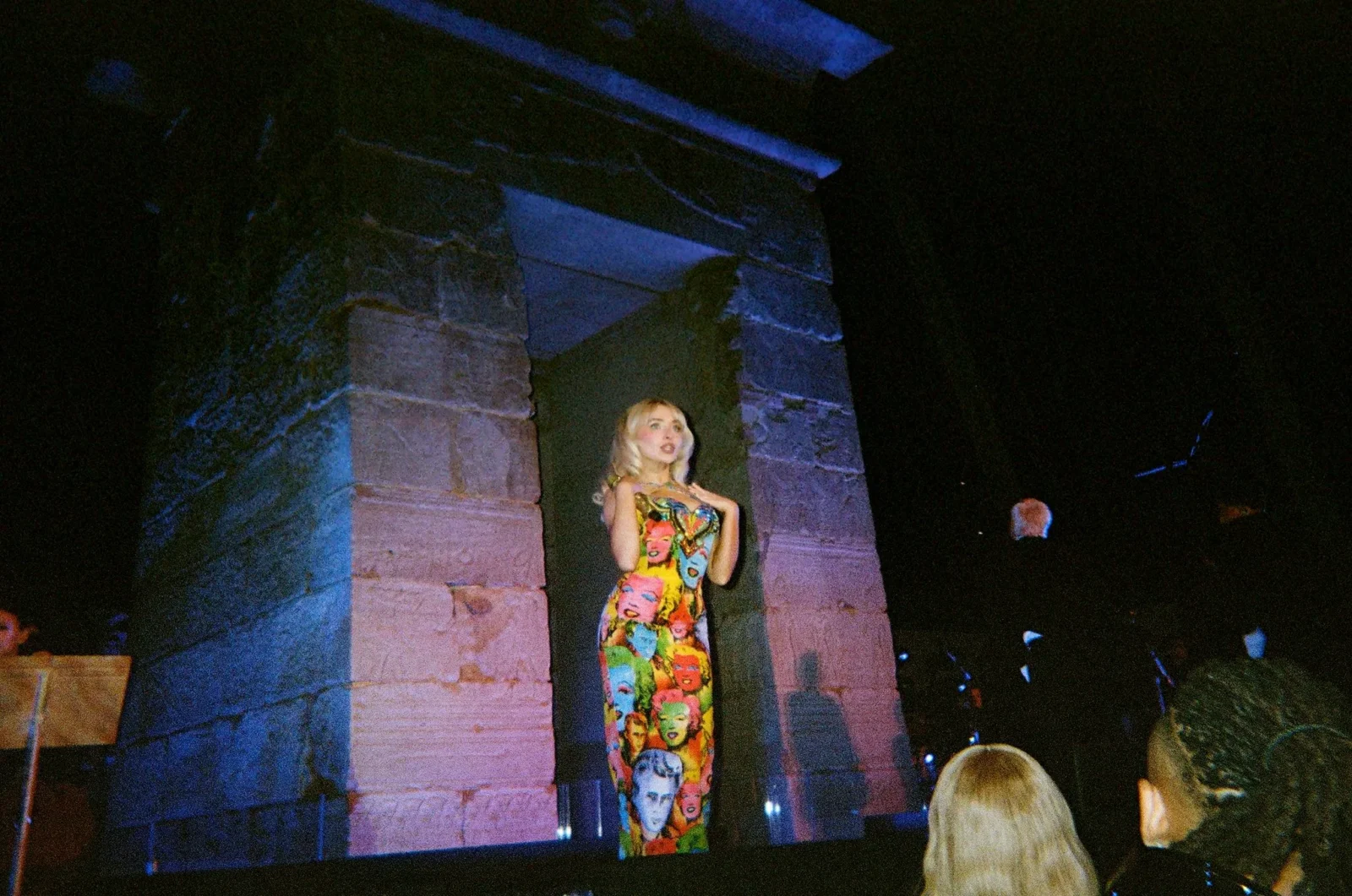 Performer in a colorful pop-art dress stands on a stone stage at night, audience in the foreground.