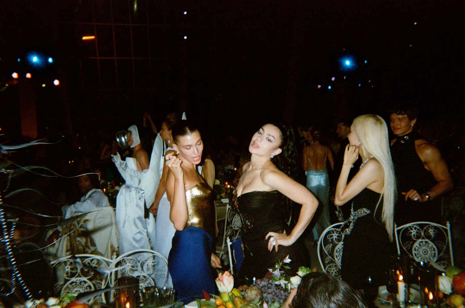 People in formal attire dancing at a nighttime party, with a table of fruit in the foreground and decorative chairs visible on the left and right.