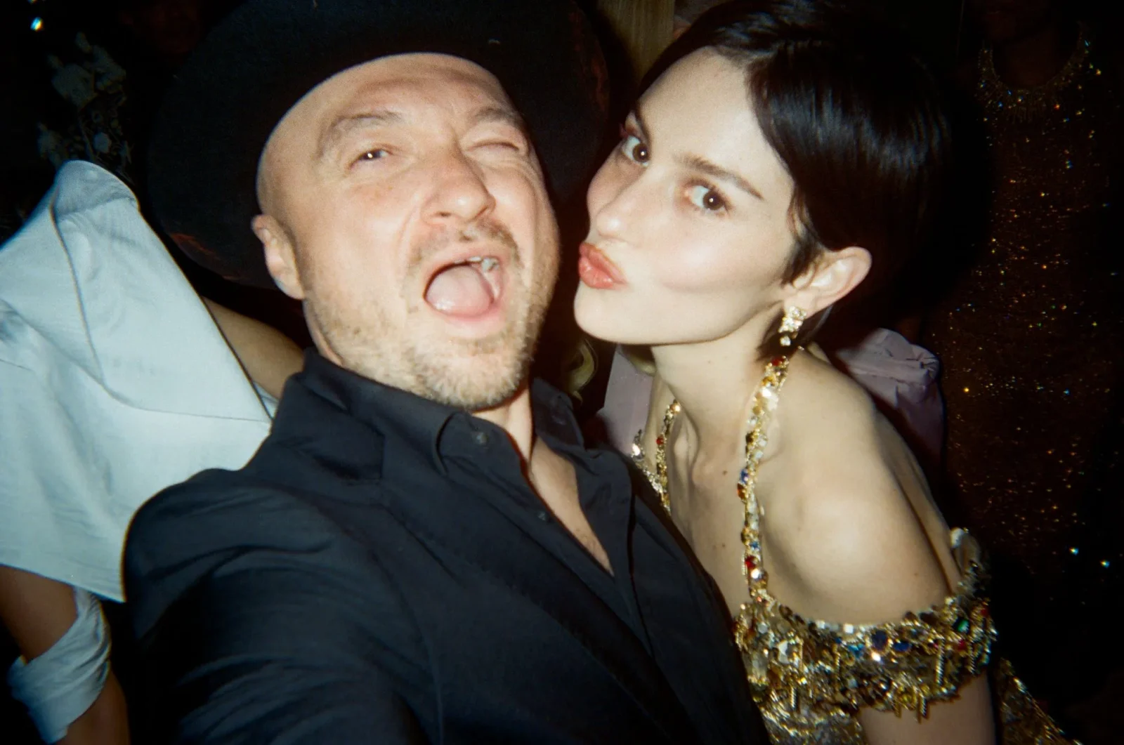 Close-up selfie of a man sticking out his tongue next to a woman in a sparkling gold dress who is kissing his cheek at a party.