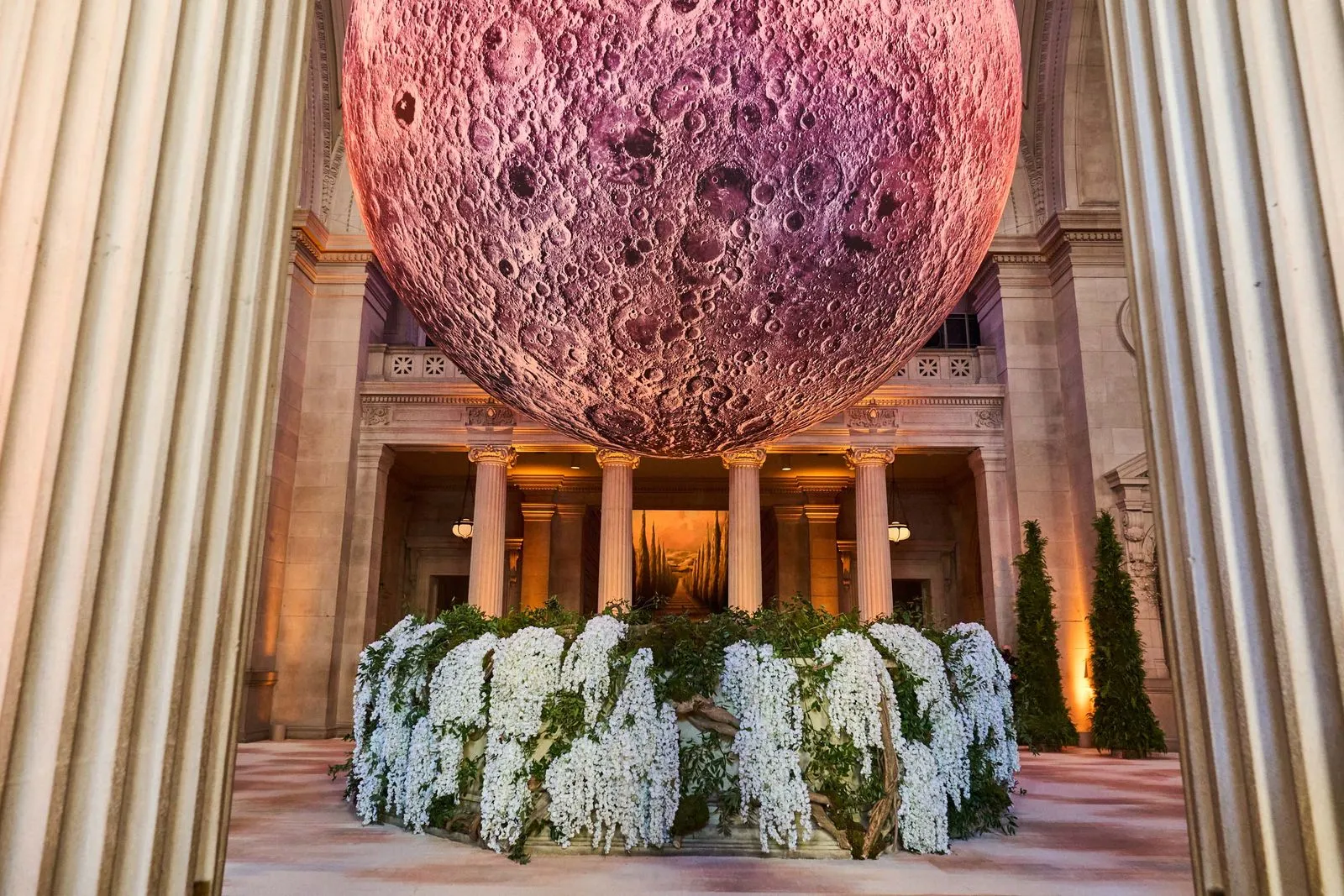 Large cratered moon sphere hanging from a grand neoclassical atrium, over a circular bed of white flowers.