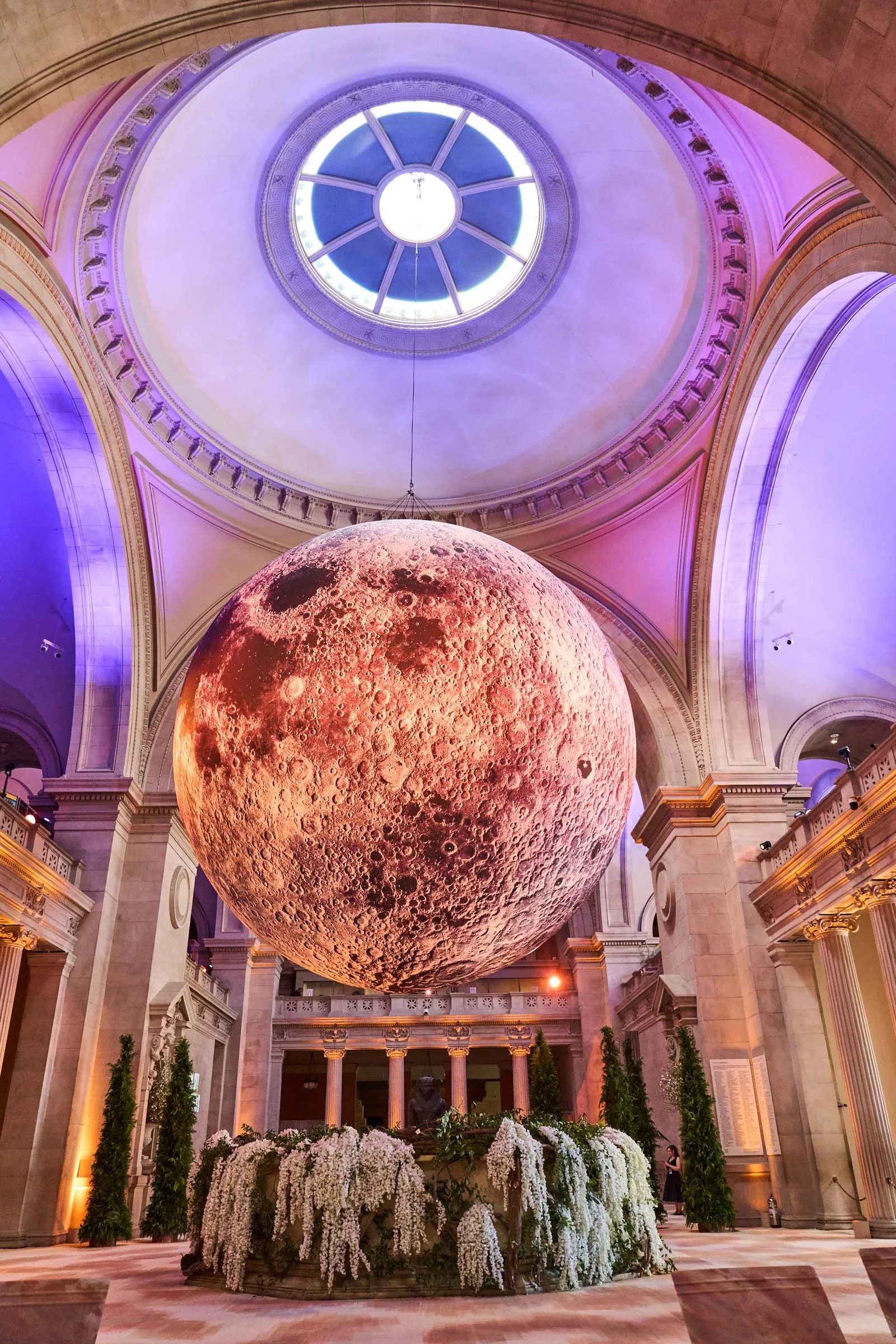 Large spherical moon sculpture suspended in a grand neoclassical hall with a circular skylight, illuminated in pink and purple hues, surrounded by cascading white flowers and plants.