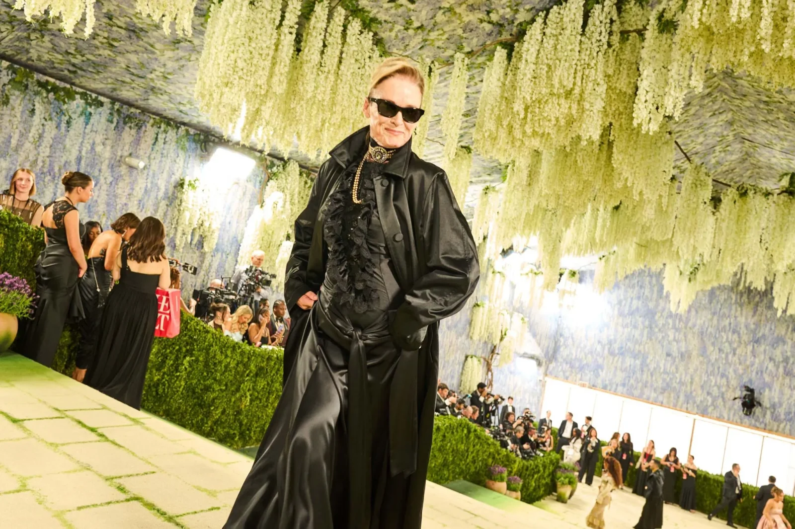 Model wearing a black satin ensemble and sunglasses poses on a runway beneath cascading white flowers at a fashion show.