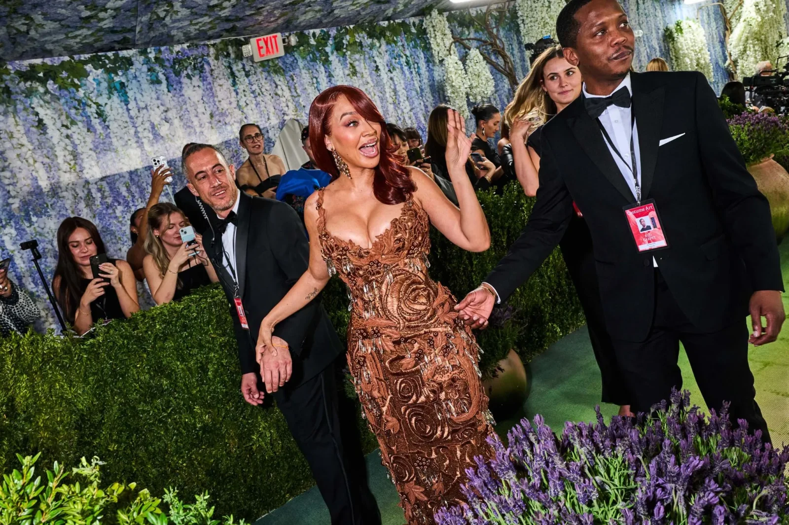Two celebrities walk a floral red carpet, woman in a bronze lace gown raising her hand as photographers film around her