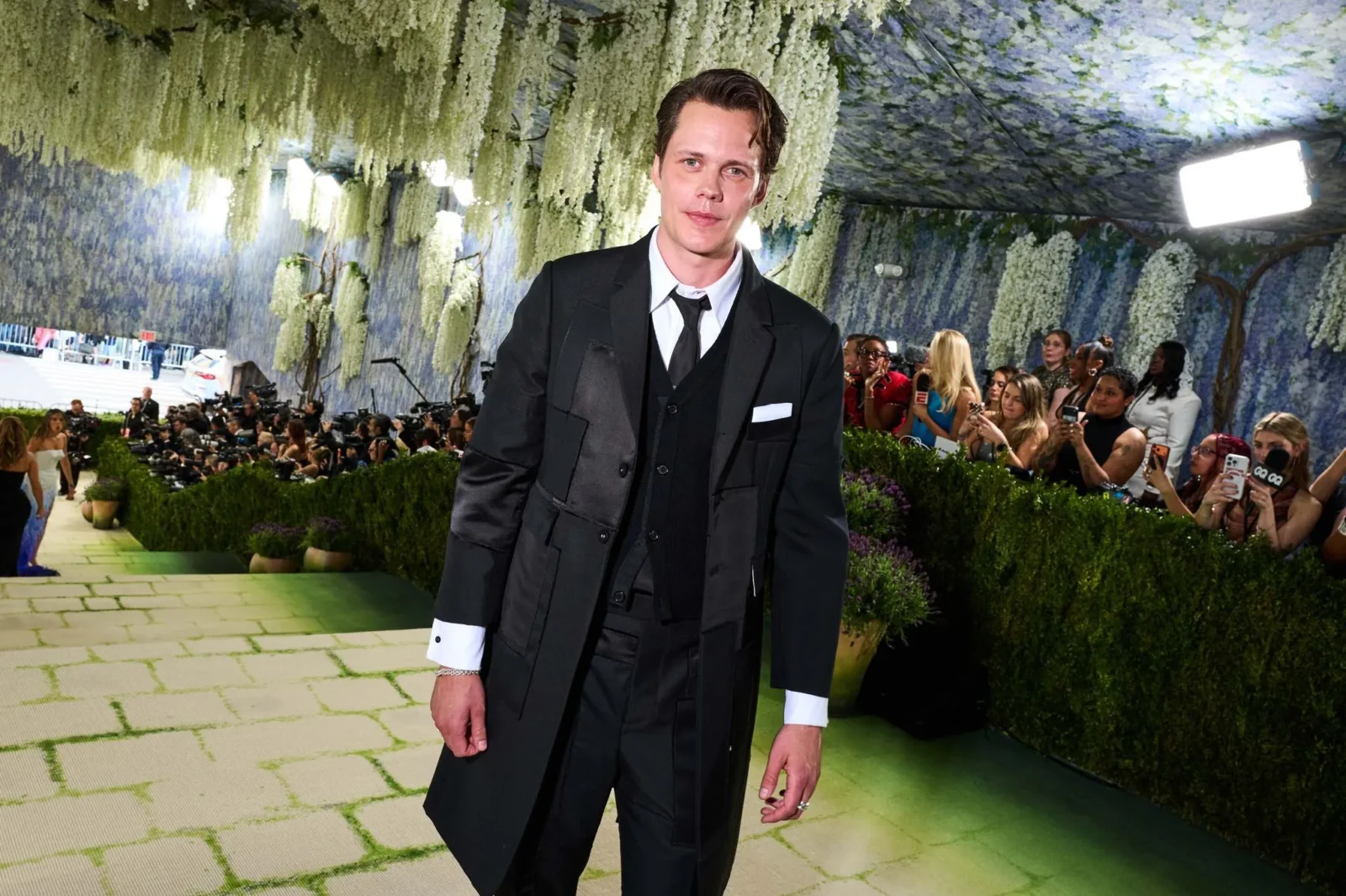 Man in a black suit walking on a green carpet at a formal event, photographers and a floral backdrop in the background
