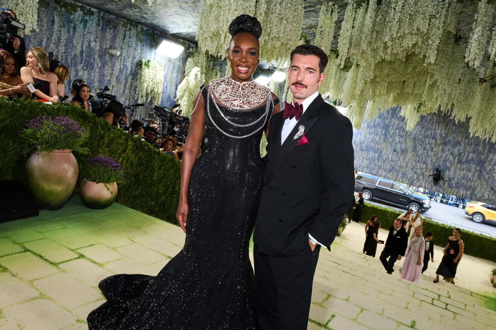 A glamorous woman in a black, sequined gown and a man in a tuxedo pose on a green-carpeted red carpet beneath cascading white flowers, with photographers in the background.
