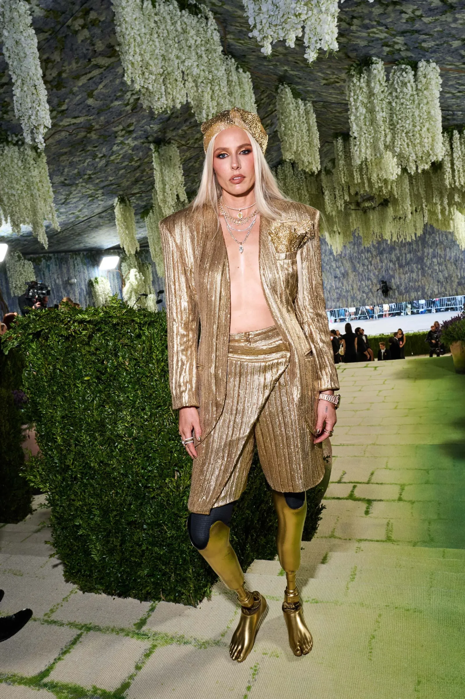 Model wearing a shiny gold pantsuit and headband, standing beneath cascading white flowers at a fashion show runway.
