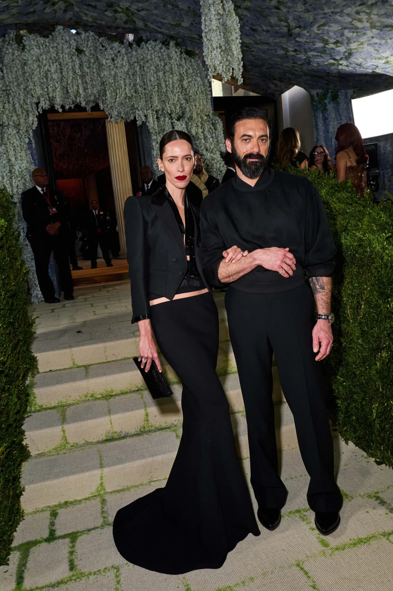 Couple posing on a staircase decorated with white flowers at a formal event; woman in a black blazer and cropped top, man in black attire with a beard.