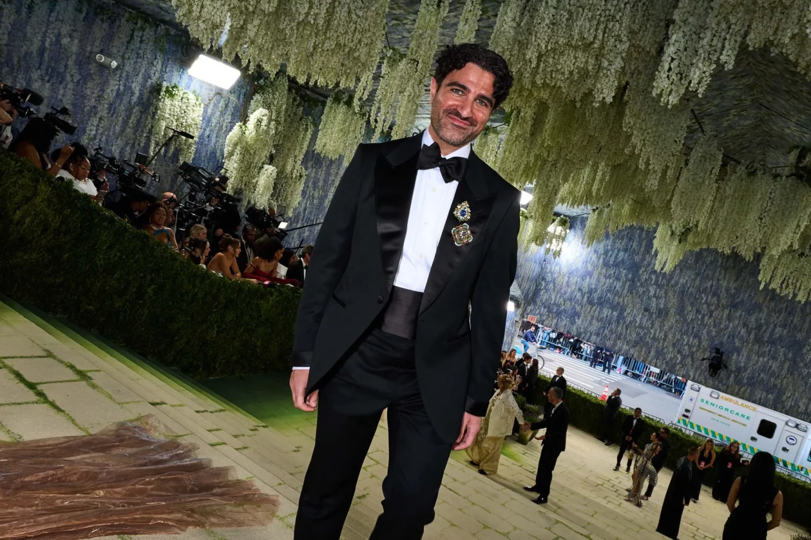 Man in a black tuxedo with bow tie posing on a green carpet at a gala, photographers and cameras in the background under hanging greenery.