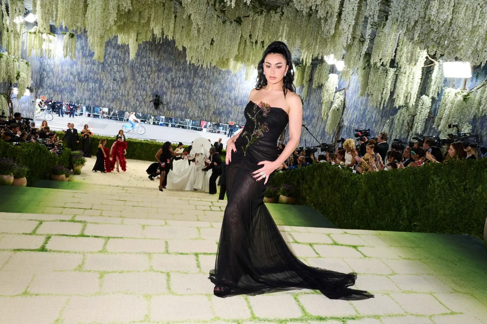 Model in a black gown walks the runway beneath hanging white flowers at a fashion show, with photographers along the sides.