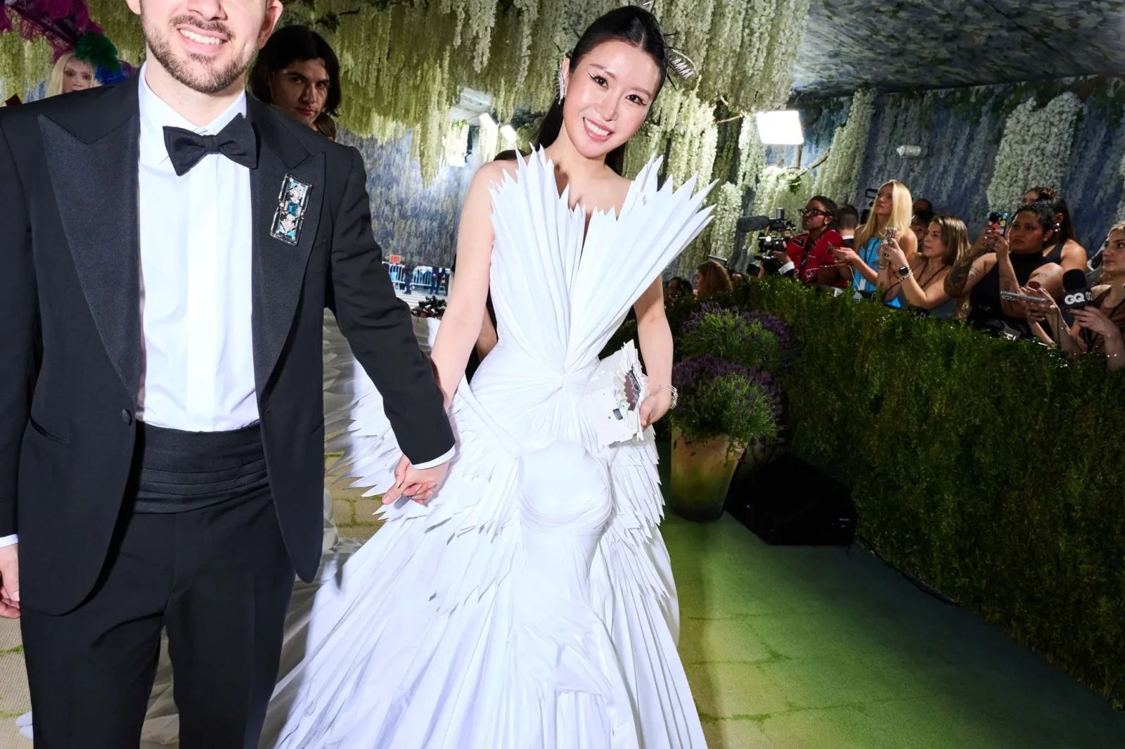 Man in a black tuxedo and bow tie holds hands with a smiling woman in a white feathered gown at a gala, photographers in the background.