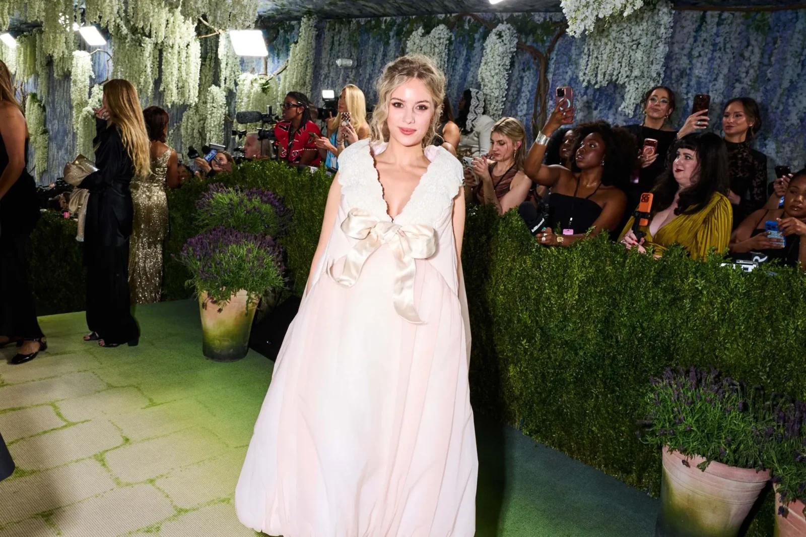 Model in a pale pink lace-trim gown poses on a green carpet at a floral-walled fashion event, surrounded by photographers and fans.