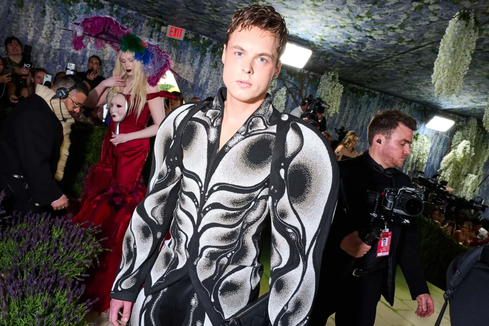 Model wearing a bold black-and-white swirling-pattern outfit walks the runway while photographers and guests crowd around amid floral decor.
