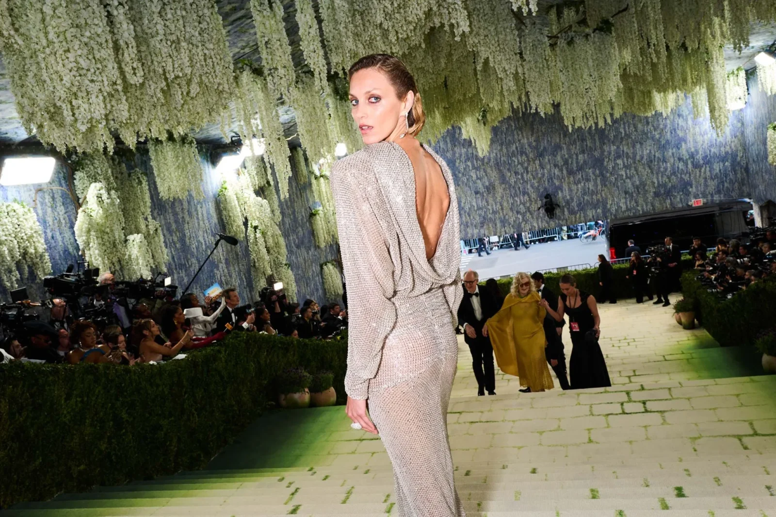 Model in a shimmering backless gown walks a runway beneath hanging white floral installations at a fashion show, with photographers in the background.