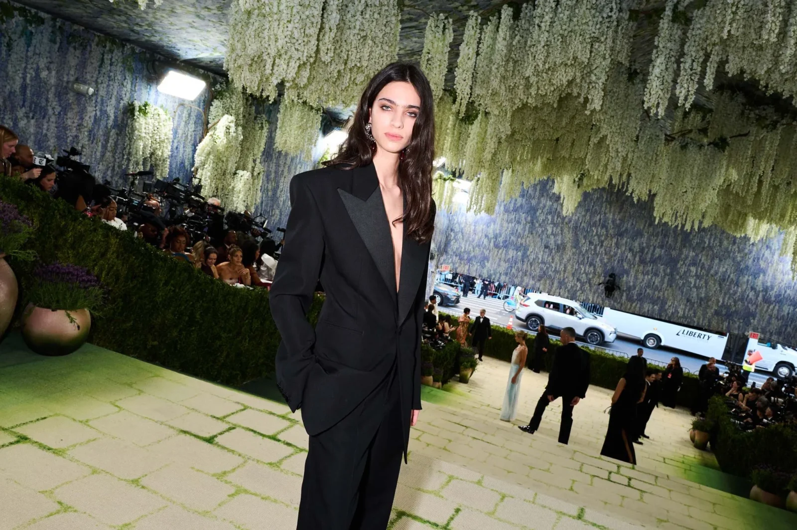 Model in a black blazer on a floral-covered runway while photographers line the left side of a fashion show.