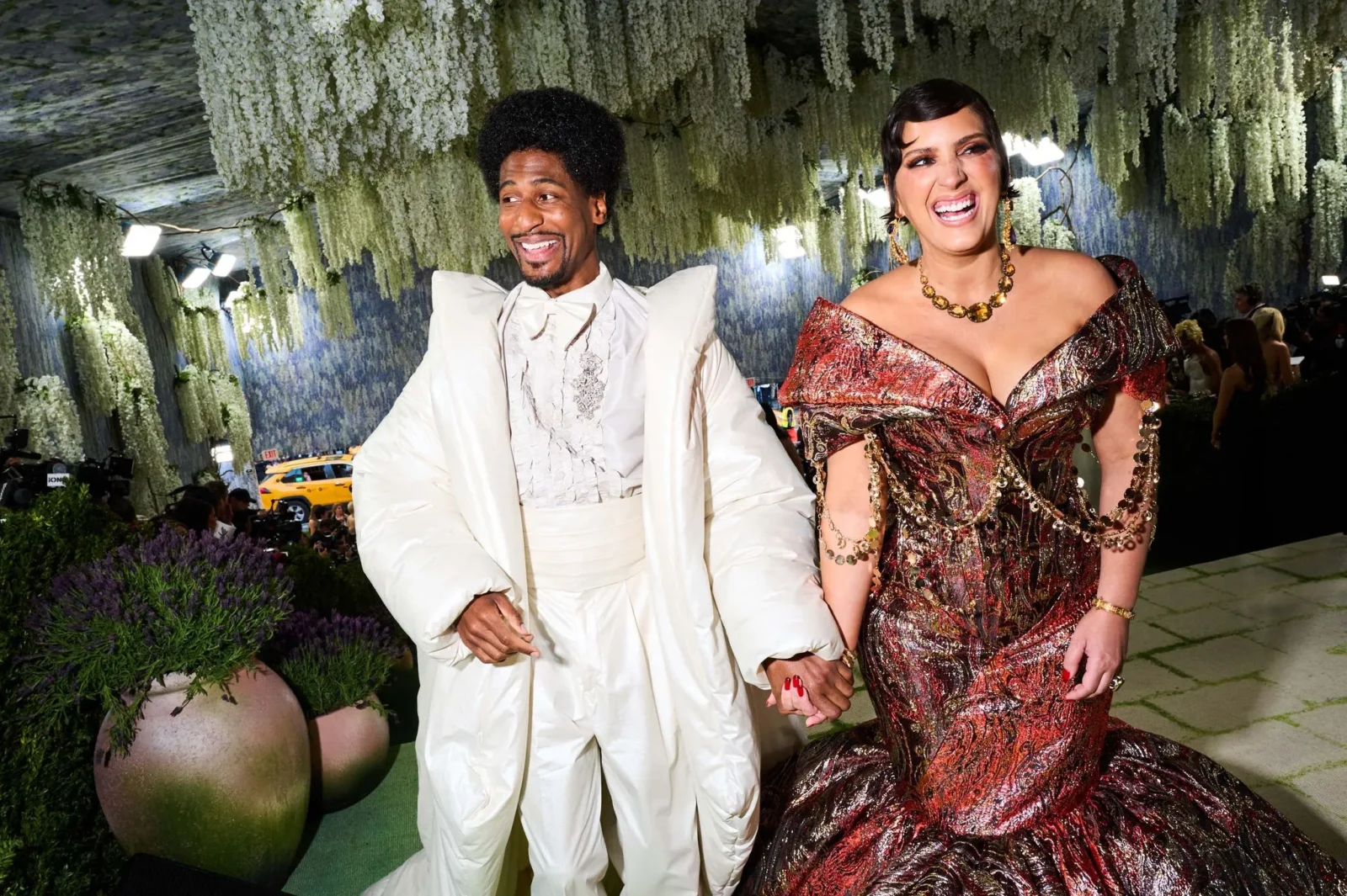 Joyful couple in formal attire walk hand in hand at a floral gala beneath cascading white blossoms above them.