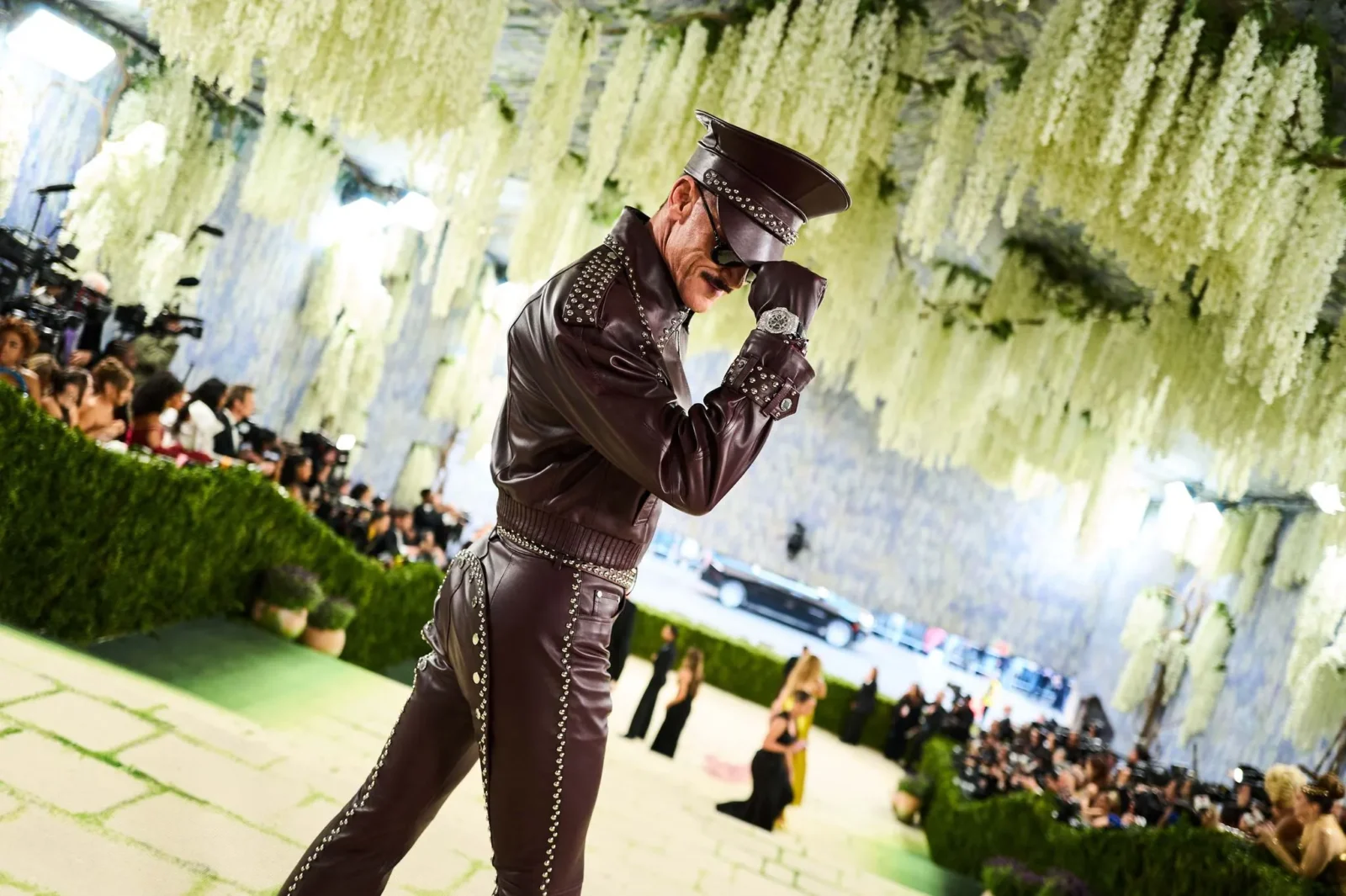 Male model in a studded brown leather outfit and cap posing on a fashion runway with cascading white flowers overhead.
