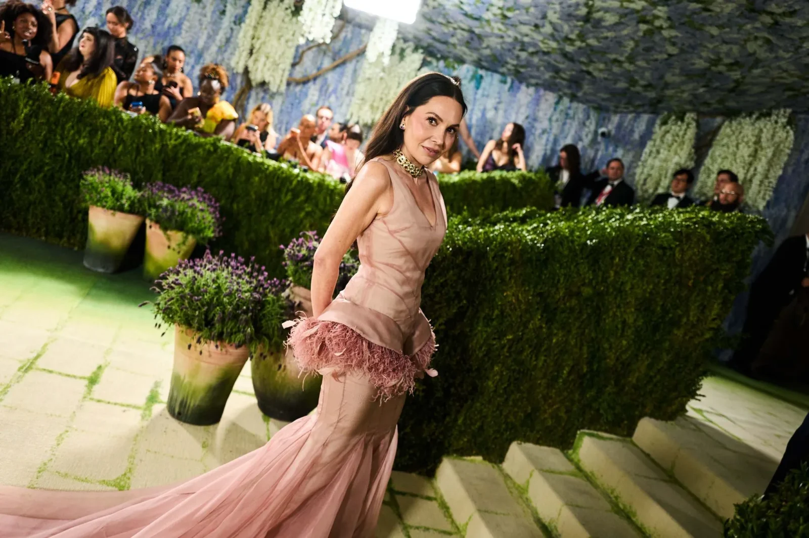 Woman in a pink evening gown walking on a runway lined with hedges at a fashion show, audience in the background watching the event