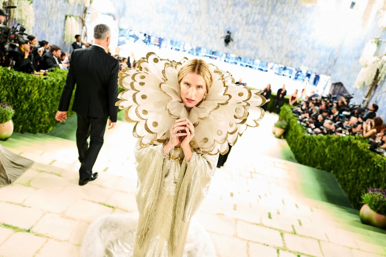 Person in an elaborate gold gown with a large ruffled collar posing for cameras at a red carpet event, photographers in the background