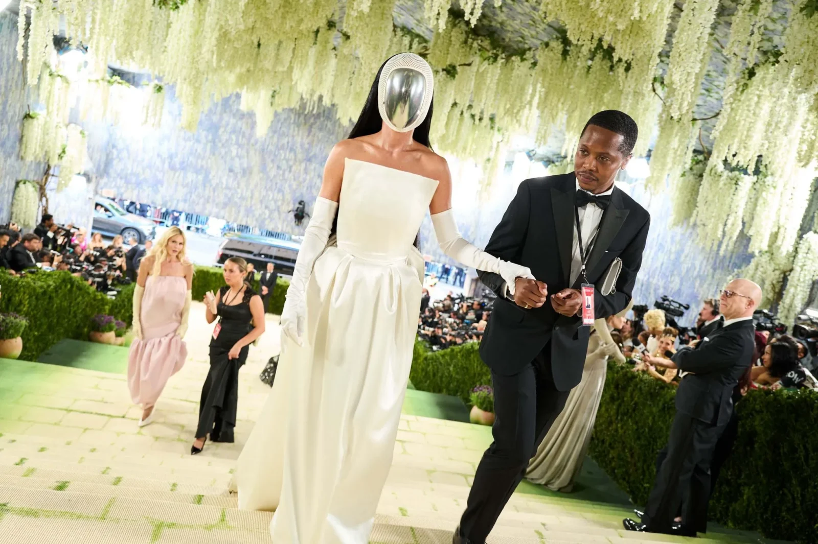 Model in a white gown and a futuristic metal mask walks with a suited escort at a high-fashion event under hanging white flowers.
