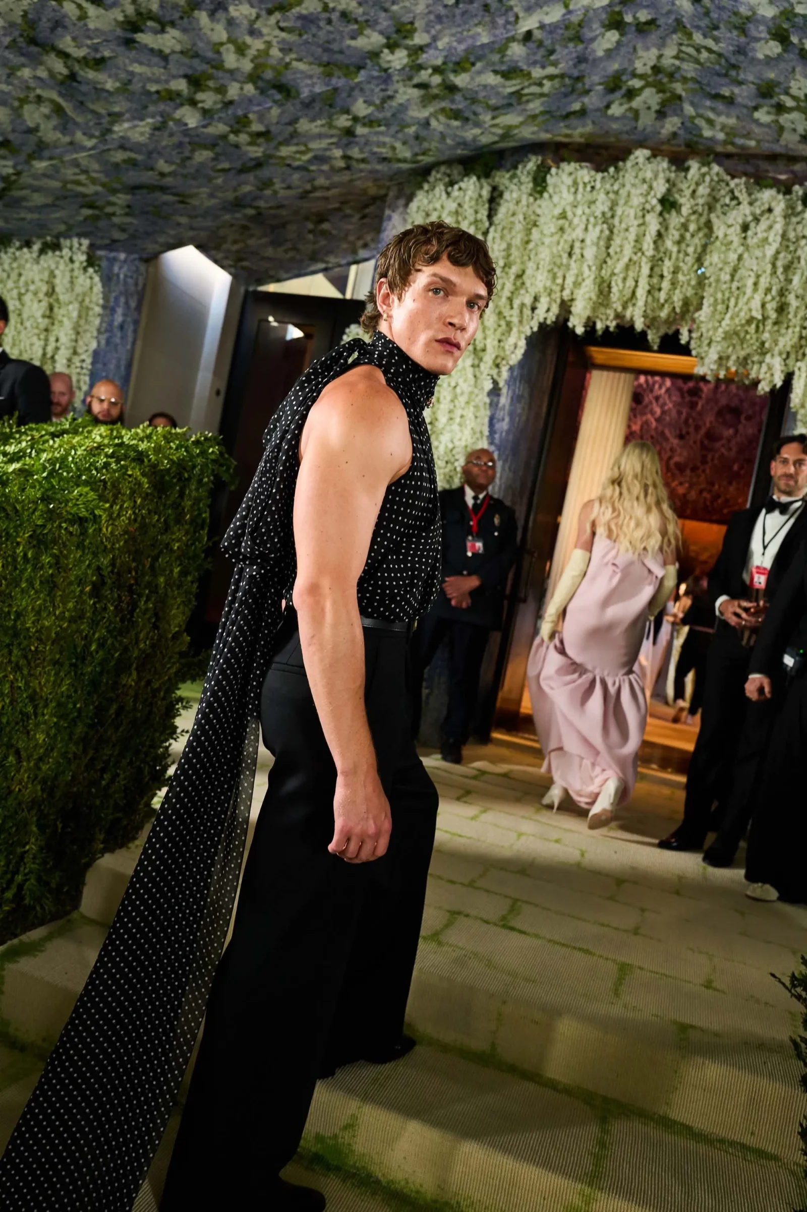 Man in a black polka-dot sleeveless top and black pants walks past a hedge at a formal event, backdrop of hanging white flowers and guests in evening wear.