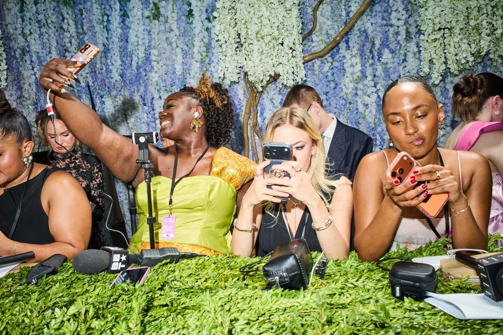 Group of stylishly dressed people taking selfies at a press event, phones raised amid microphone gear and a floral backdrop.