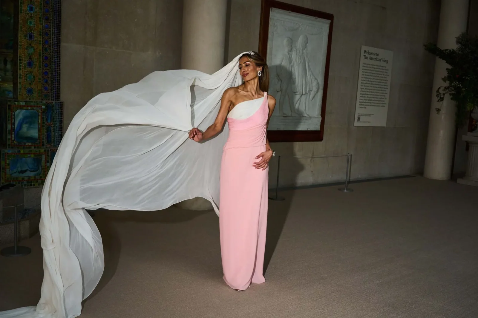 Woman in a pink one-shoulder gown with a long white cape sweeping behind her, in a museum hallway.