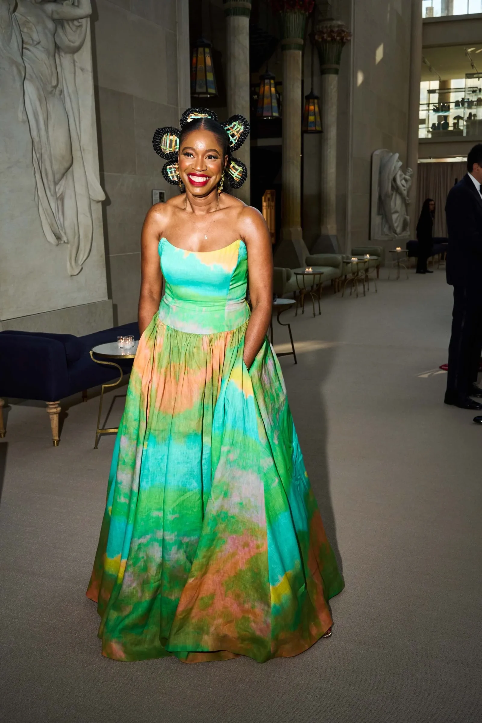 Smiling woman in a colorful turquoise and green gown with braided circular hair accessories, standing in a marble lobby with statues in the background.