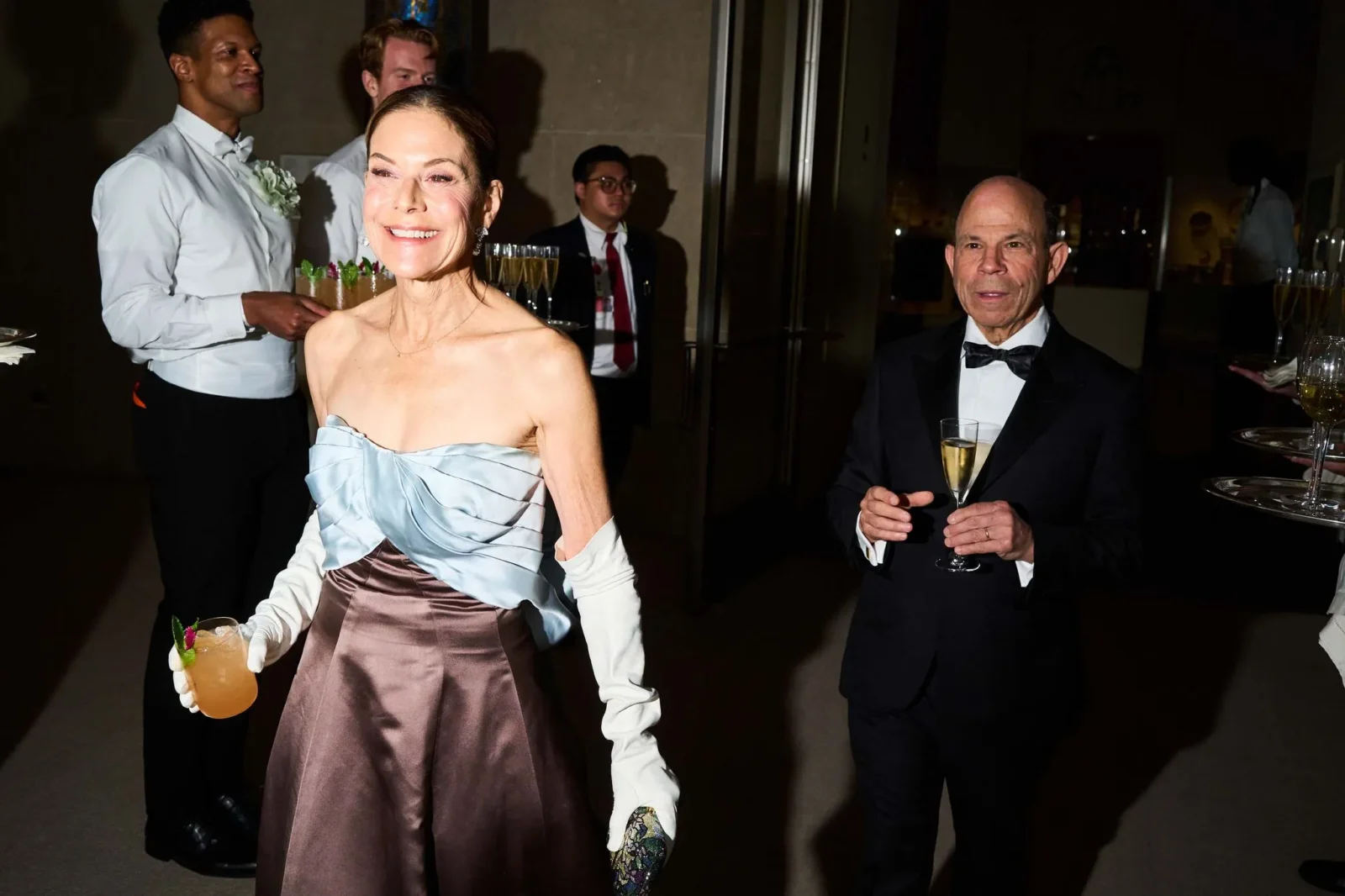 Smiling woman in a blue-toned gown and gloves holds an orange cocktail beside a man in a tuxedo at a formal party.