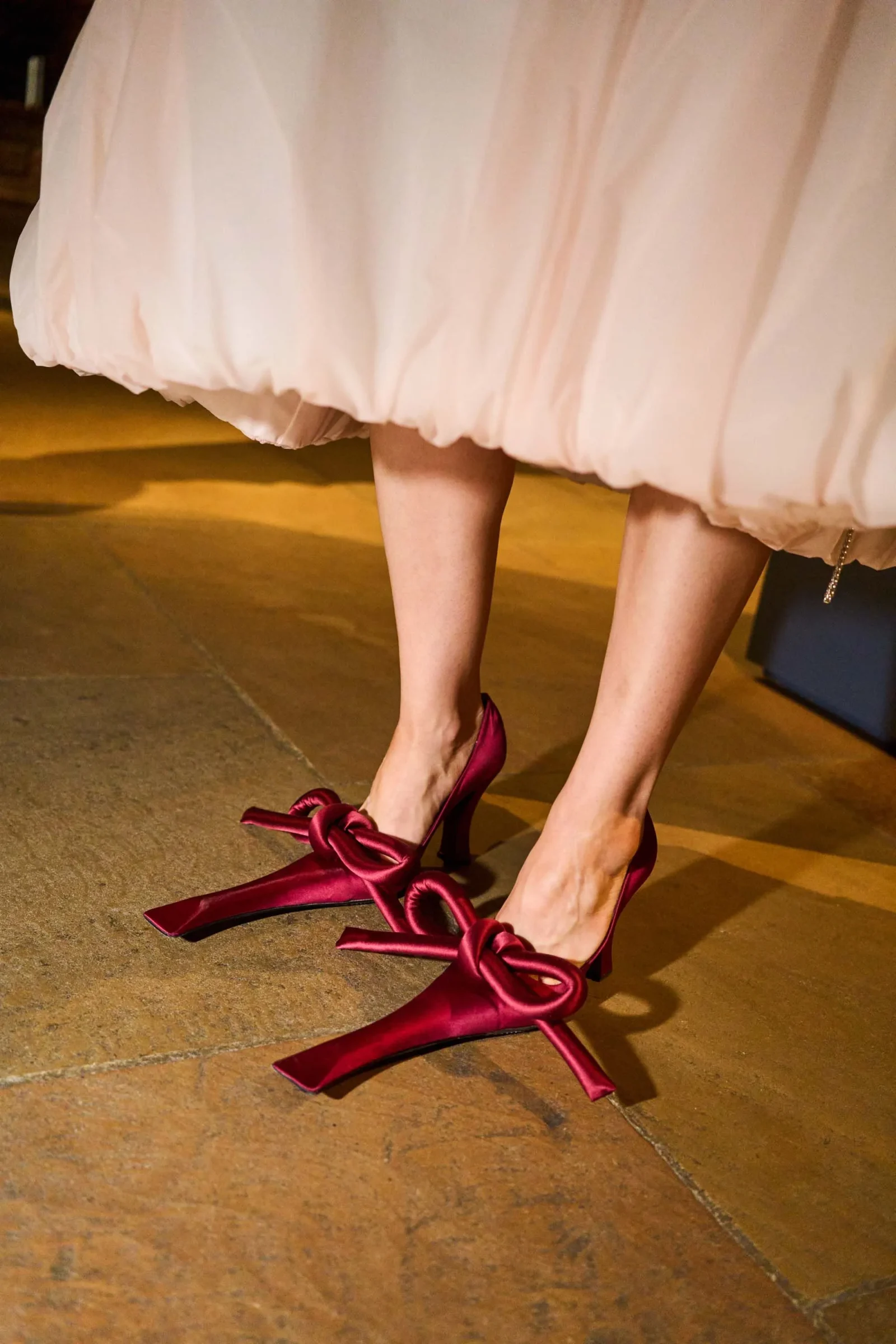 Close-up of a pale pink, voluminous dress hem and burgundy high heels with large bows, standing on a warm-toned floor.