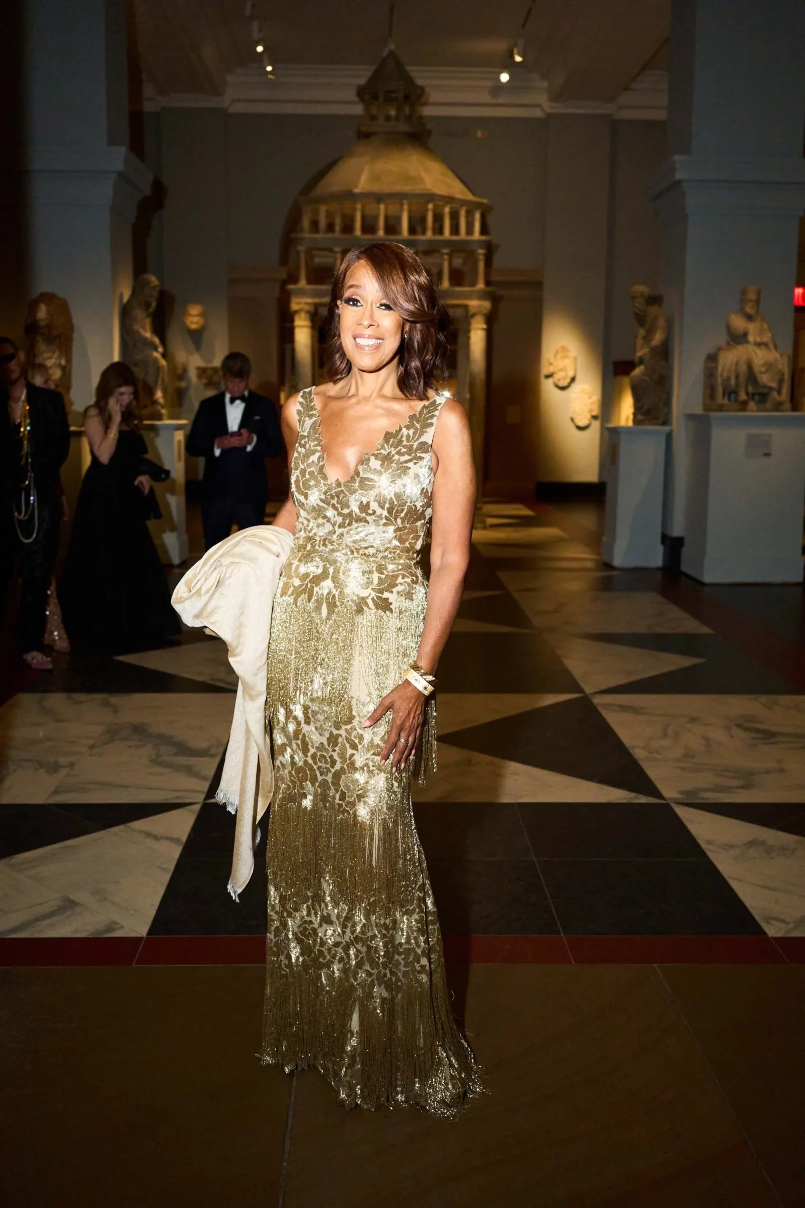 Smiling woman in a sparkling gold lace gown holding a light wrap, posing in a museum gallery with classical statues behind her.