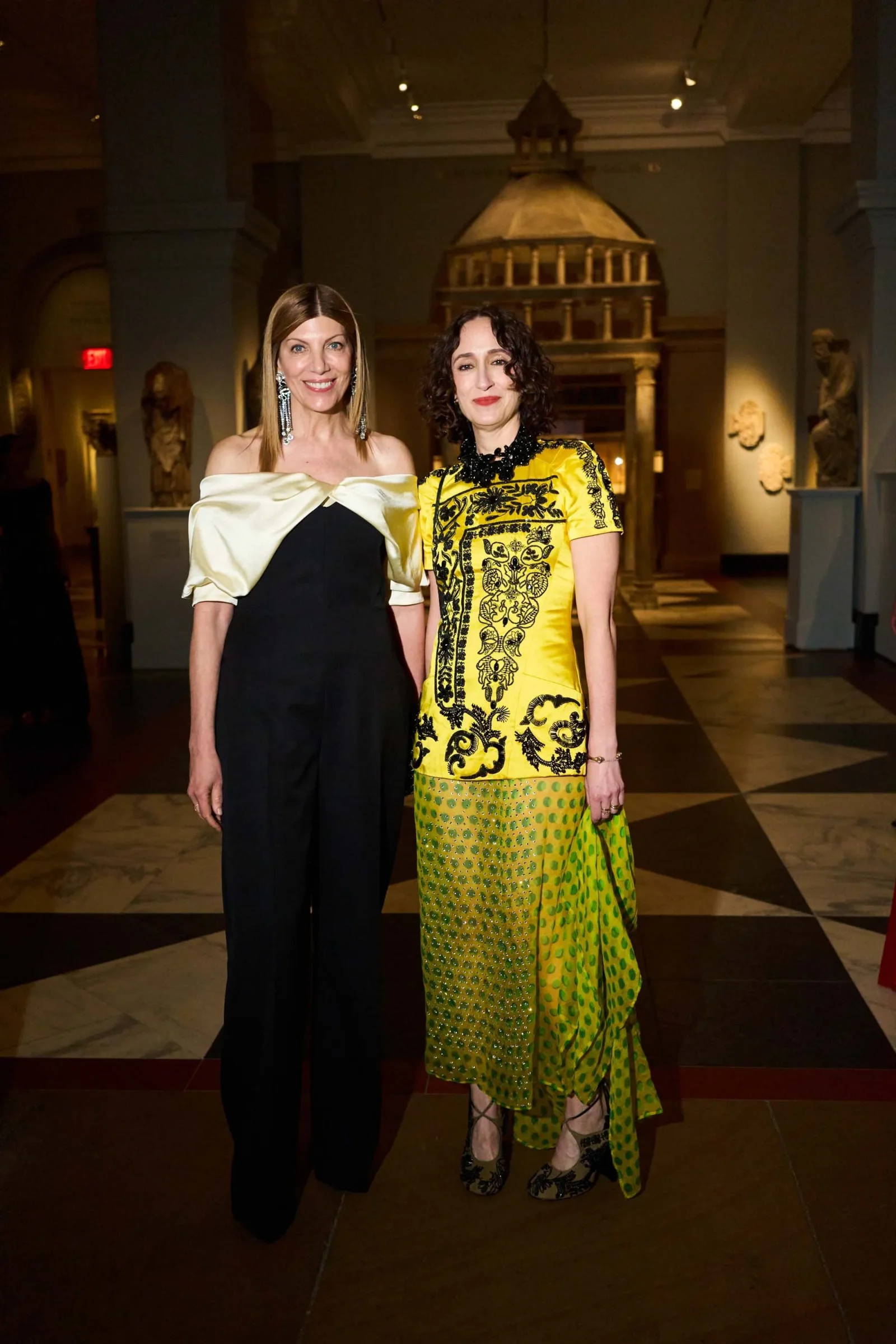 Two women pose together in a museum gallery, smiling; one wears a black and white off-shoulder dress, the other a yellow and black patterned gown with green accents.