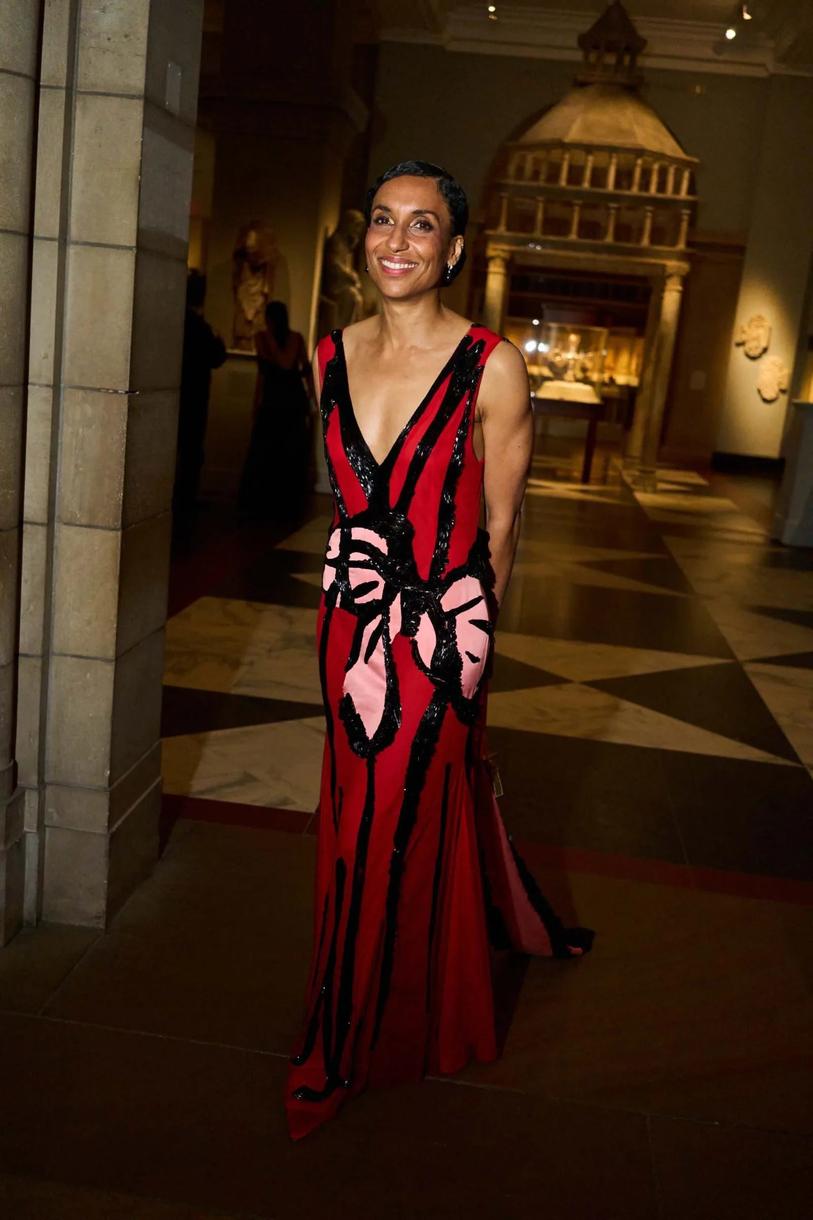 Smiling woman in a red and black patterned evening gown stands in a museum gallery with statues and display cases in the background.