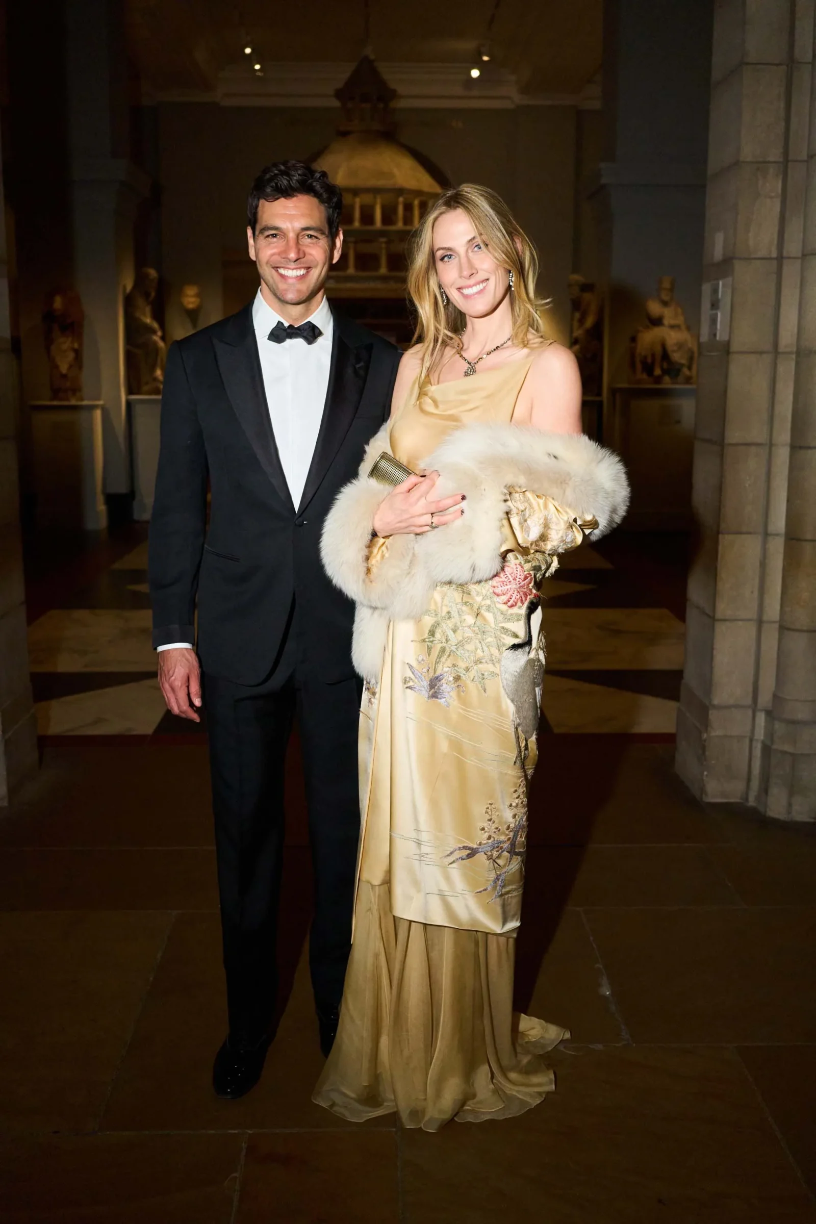 Man in a black tuxedo and bow tie stands beside a smiling woman in a gold gown with a white fur stole at a formal event, museum backdrop behind them.