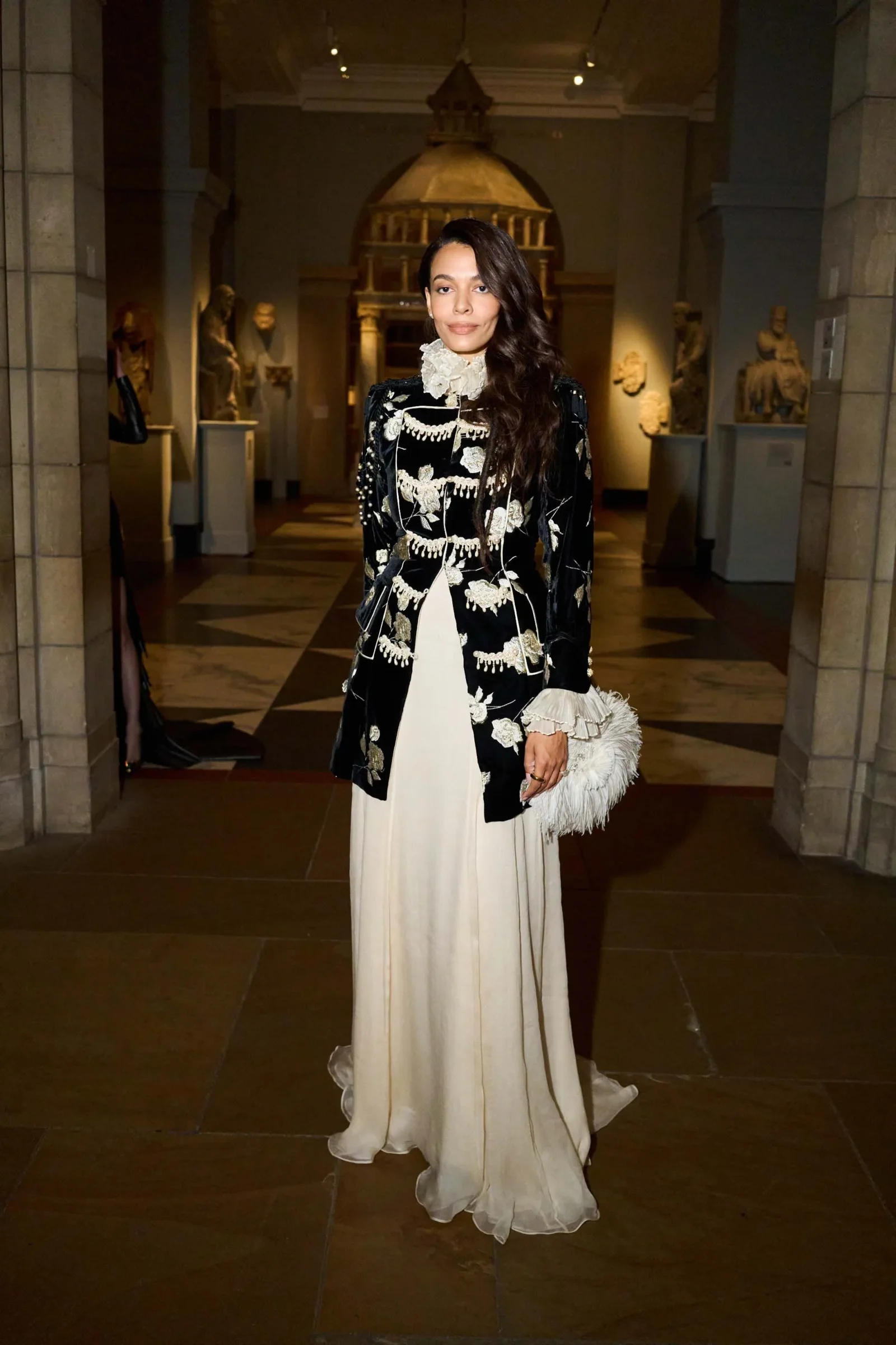 Woman in a black-and-white embroidered coat over a cream gown posing in a museum gallery with statues in the background.