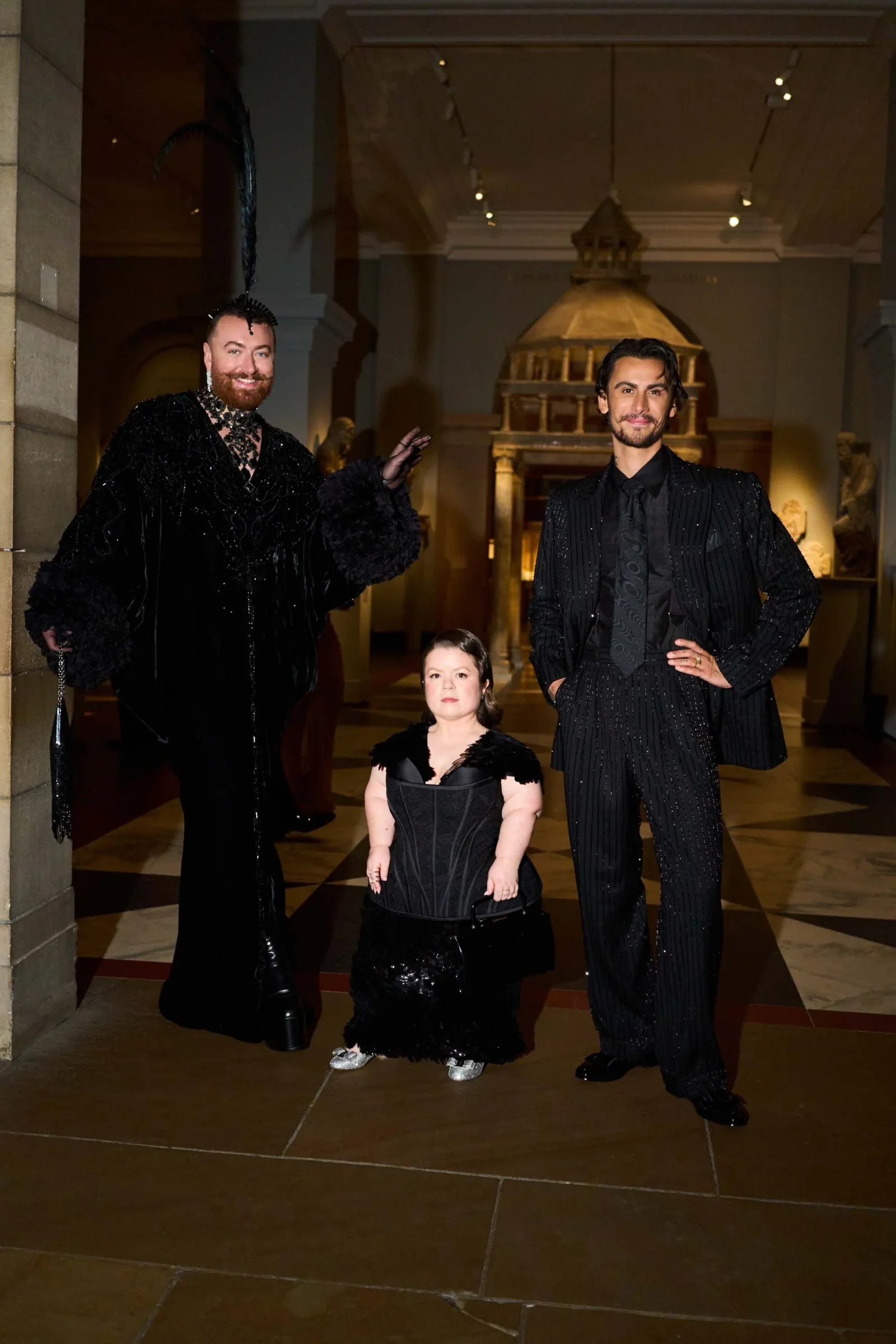 Three people in black formal outfits pose in a museum hall with marble columns behind them, two tall men flanking a child in the center.