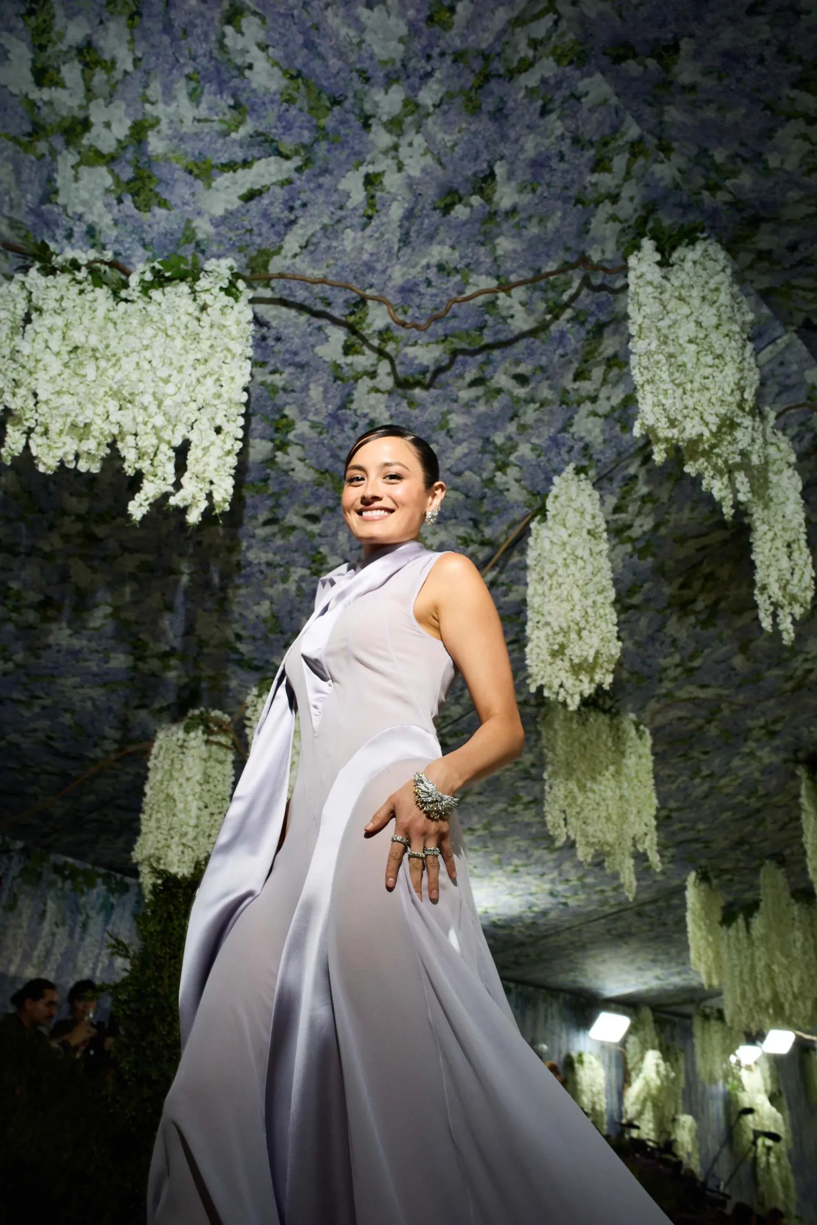 Smiling woman in a silver satin gown walking on a floral runway with cascading white blossoms overhead.