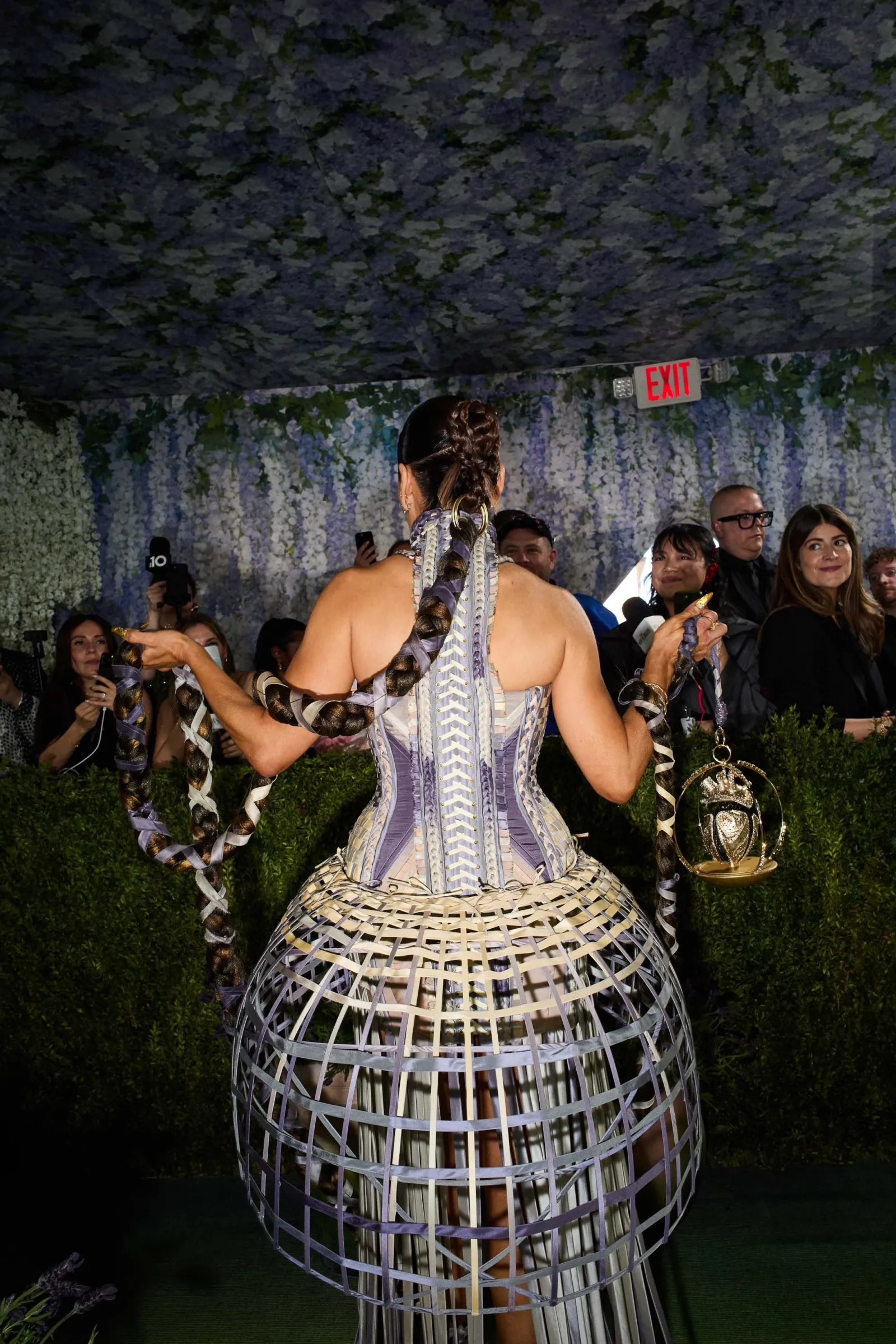 Person wearing an intricate cage-like dress with braided back, standing on a runway as onlookers watch in a decorated hallway with an exit sign above.